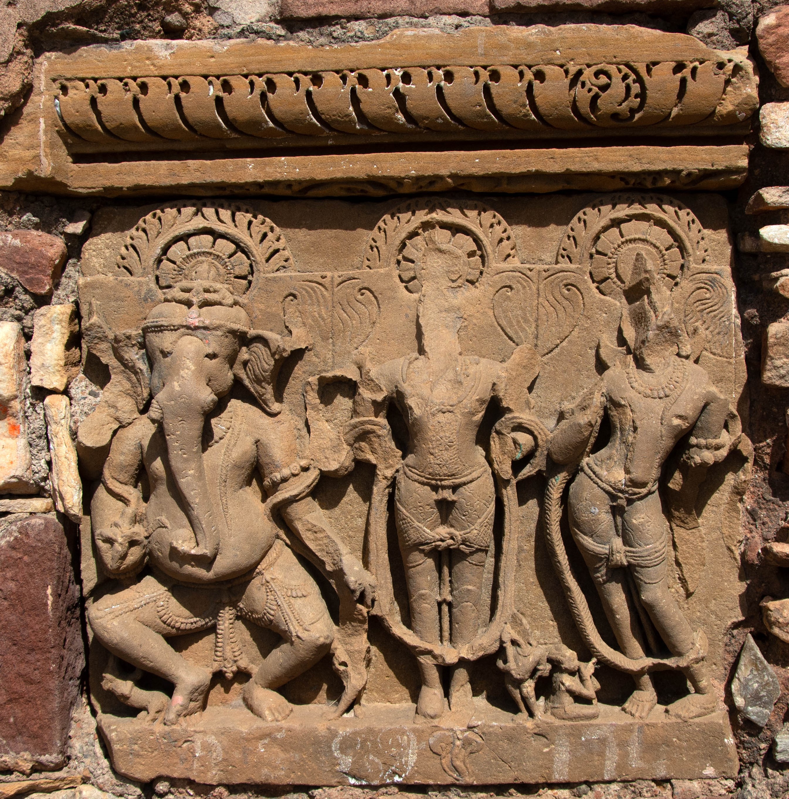 Image 5: Affixed to the later structures at the site are several loose sculptures, including the trio of Ganesha, Surya, and Chandra. The three figures may have once been part of a larger panel depicting the nine heavenly bodies, or navagraha. While the dancing Ganesha has four arms, the images of Surya and Chandra only have two, which are both broken. Surya's erect uplift posture, or samabhanga, contrasts with Ganesha's dynamic posture and Chandra's tribhanga pose.