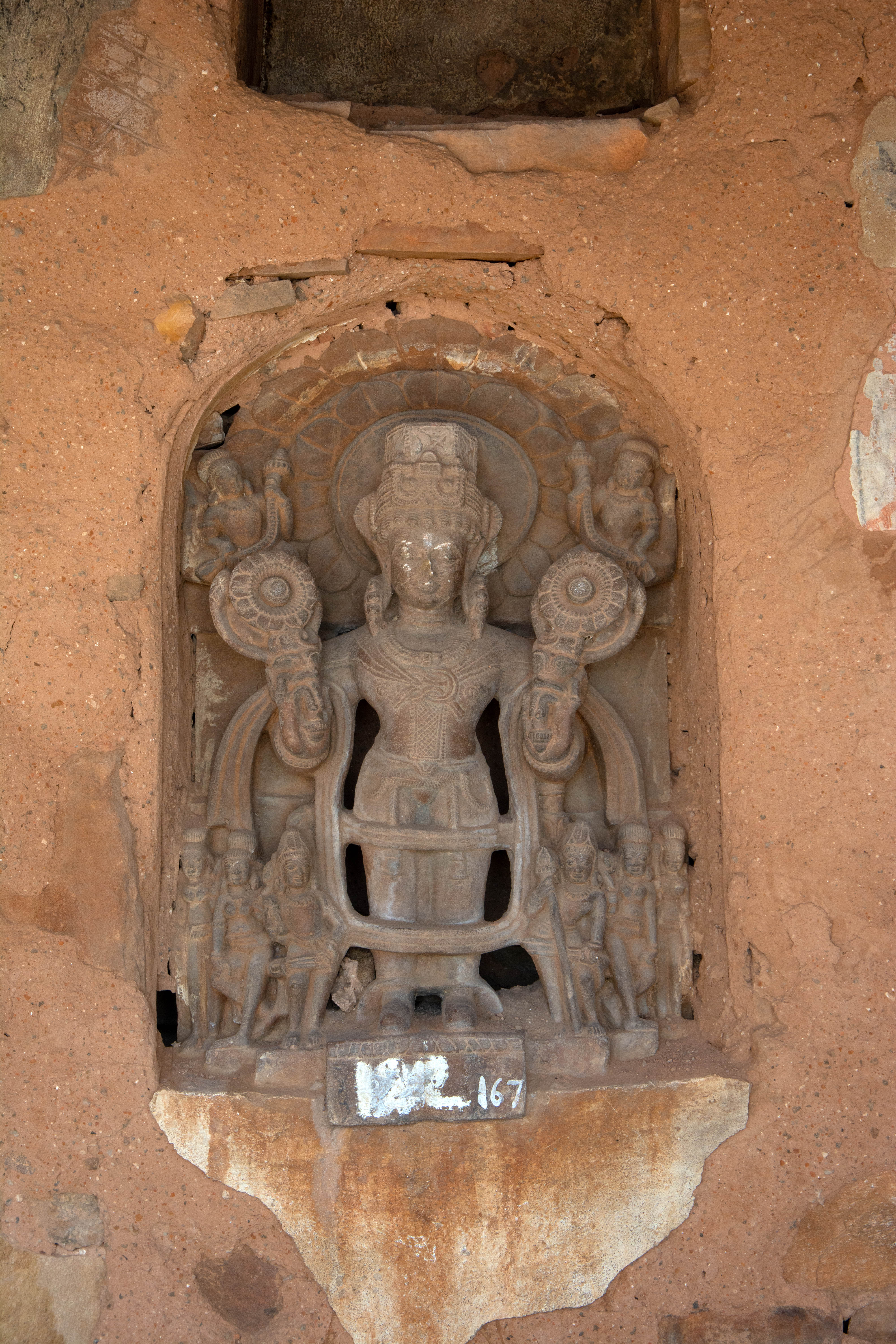 This sculpture is arguably the oldest at the shrine, dating back to the 8th century CE, according to Ambika Dhaka. Thus, the sculpture significantly predates the construction of the Harshnath Temple in the second half of the 10th century. The figure exhibits standard iconographic features of the Sun God, such as fully bloomed lotuses in two hands, a long tunic, boots, and retinue figures in pairs like his companions, Danda and Pingala, his wives, and his sons, Ashvins.