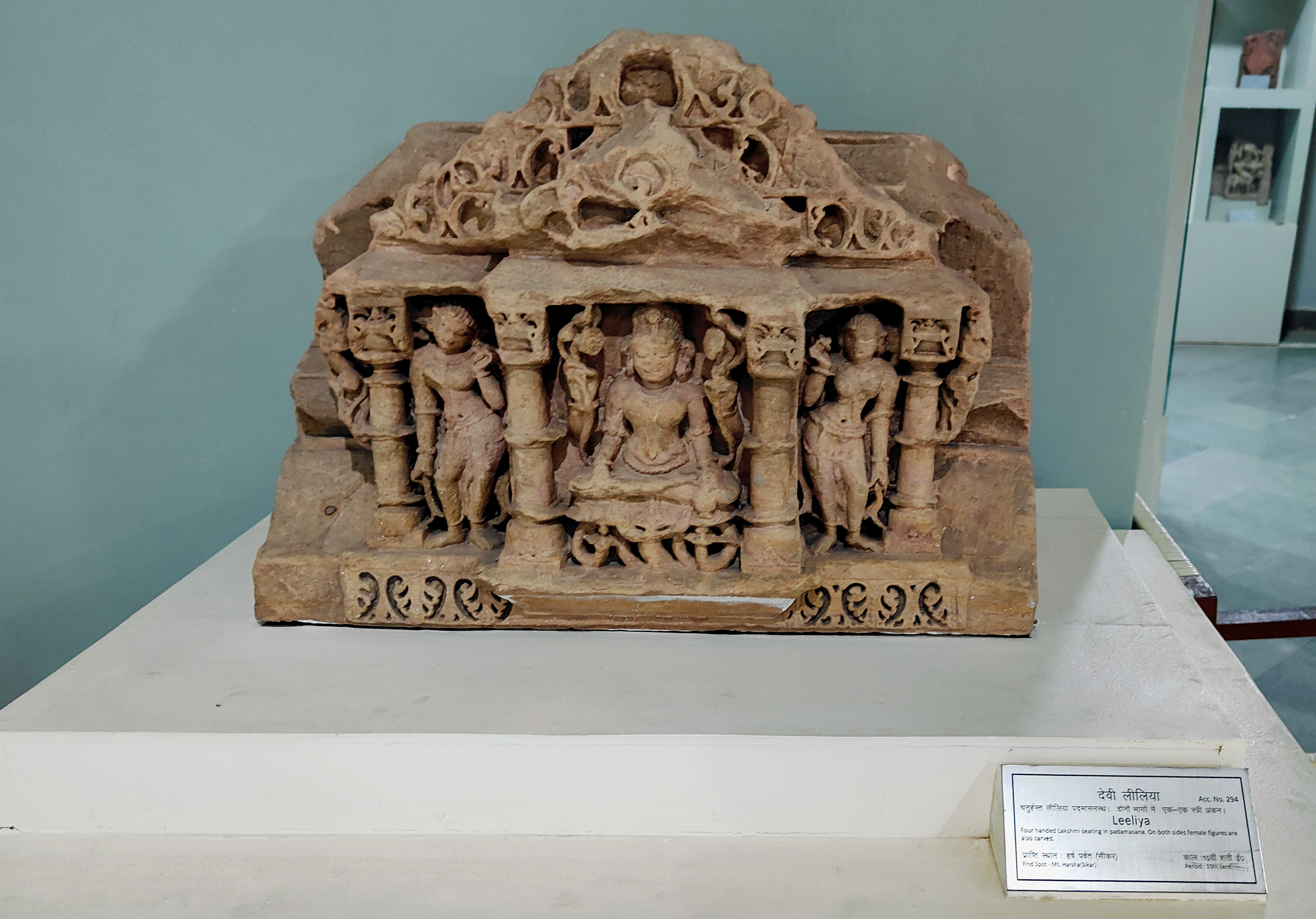 The statue depicts a seated Lakshmi, holding lotuses in her upper two hands, her lower left hand clutching a pot, and her lower right hand in the varada mudra. The museum label refers to her as Goddess Leeliya. On either side, female attendants holding flowers accompany her.