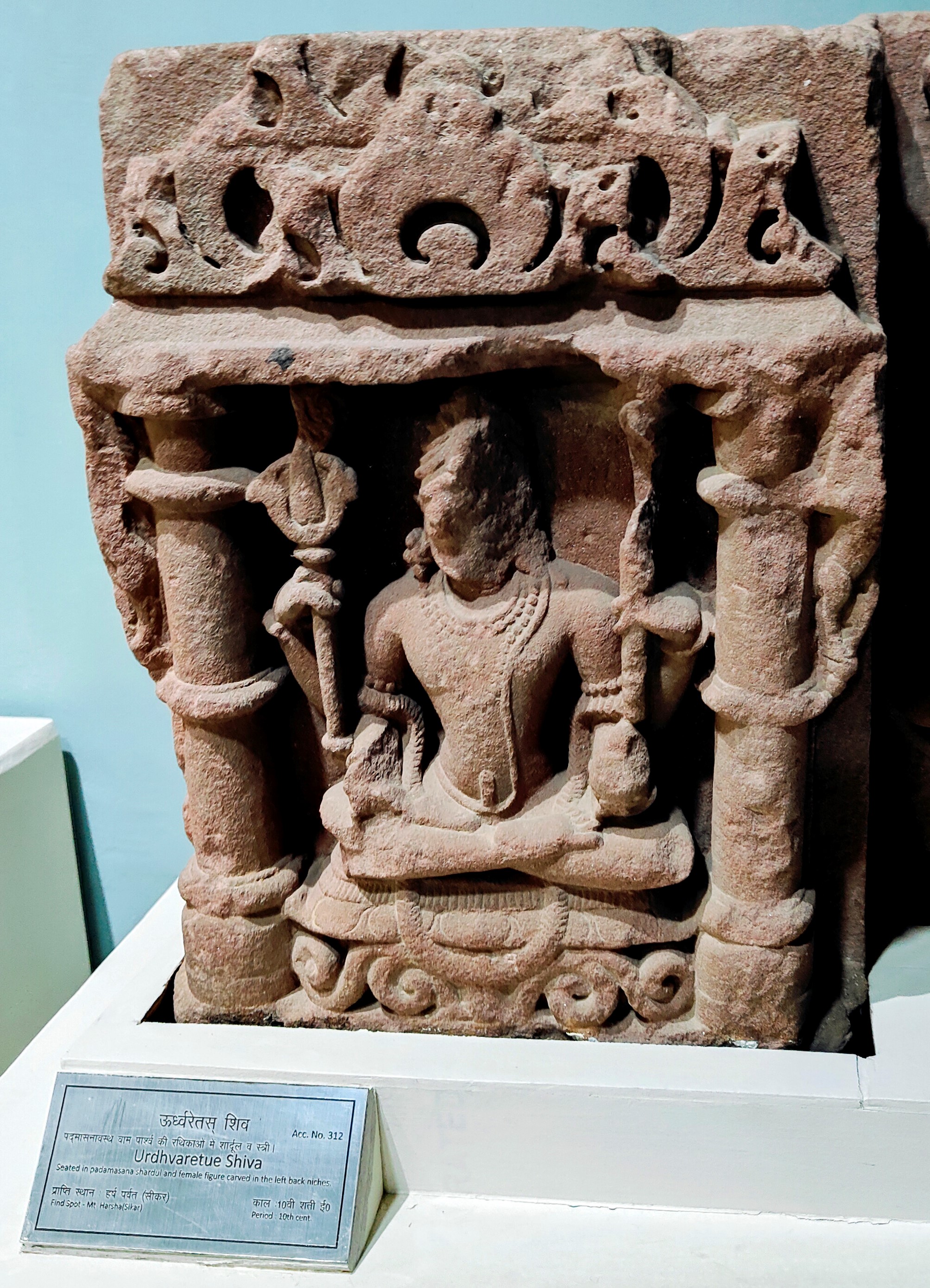 Image 13: This Shaiva figure was possibly part of a devakostha or sculptural niche placed on the walls of a now-lost Shaiva shrine. It is now housed in the Government Museum, Sikar. The four-armed seated figure holds a trident and a staff in the upper two hands, while the lower two hands are considerably damaged. The figure is ithyphallic, symbolizing the Urdhvareta aspect of Shiva, indicating mastery over the vital energies through ascetic vigour.