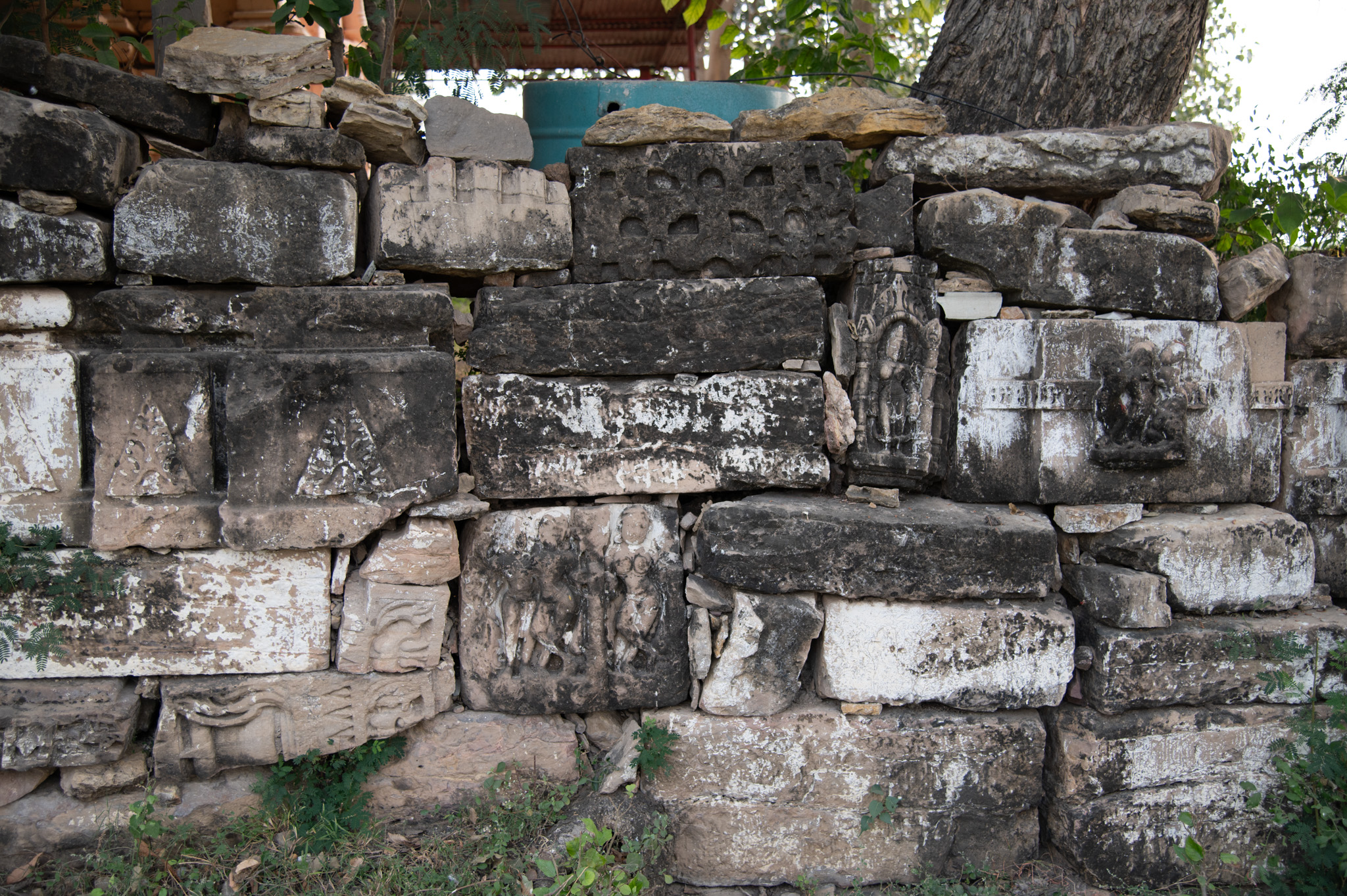 The temple's broken fragments, including the vedibandha mouldings, form a boundary wall around the temple by placing them close to each other. Figures of deities, females, and couples adorn some of the mouldings. Also visible are fragments of pillars.