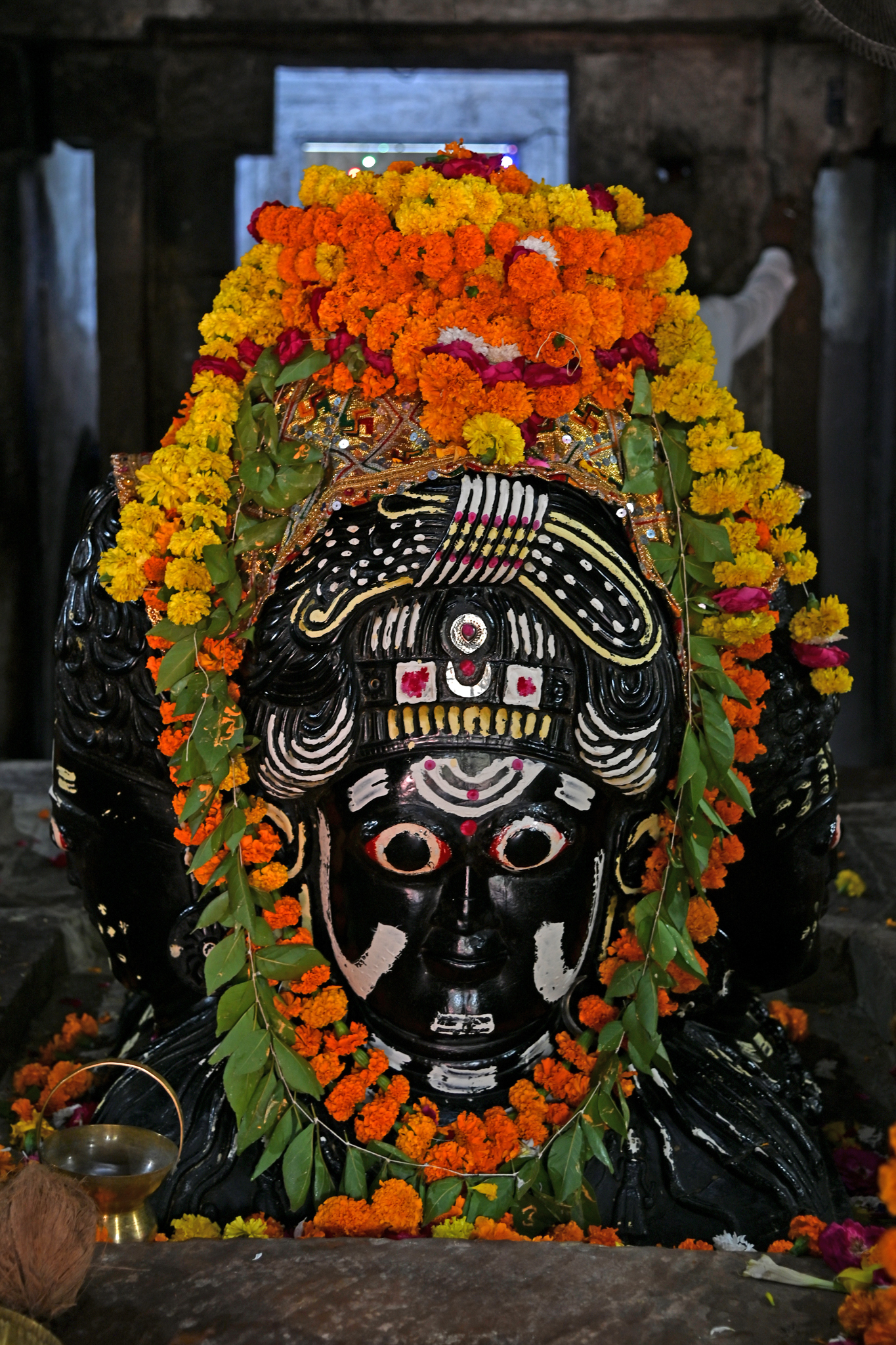 Image 7: A Chaturmukha linga is placed on top of the stone platform in the mandapa. As per the literal meaning of the term Chaturmukha, the linga has four faces, each facing a cardinal direction. The linga is made of a black stone and is worshipped as the primary deity by the locals. According to popular lore, the Chaturmukha linga represents Shiva, Parvati, Brahma, and Vishnu. The mukha facing the east (entrance gate) is of Shiva with a heavy jatabhara (braided hairdo) on the head. The south face of the linga has a heavy jatamukuta (matted crown) on its head and hairlocks cascading down to the shoulders. The west face (rear) also has a jatamukuta, albeit carved slightly differently from the other three. The north face has snail-shaped curls culminating in a jatamukuta.