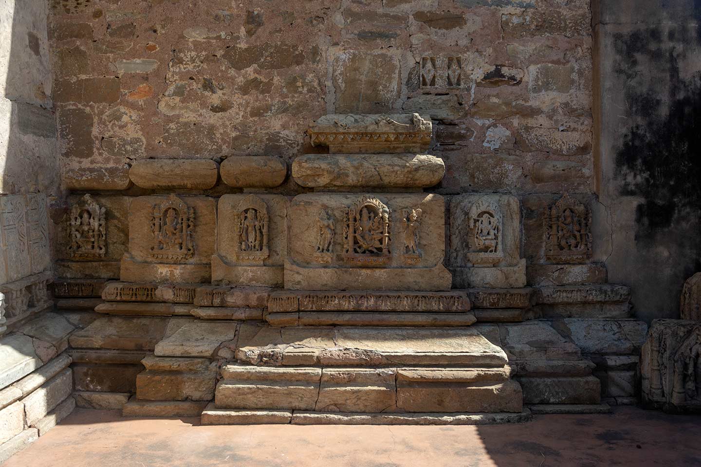 This is the exterior of the southern shrine in the Neelkanth Mahadev triple-shrine temple. The shrine's walls have fallen, leaving only the plinth up to the level of the kumbha (pot) moulding in situ. Only a few traces of the kalasha (pitcher) moulding above the kumbha have survived. The faces of the kumbha moulding have depictions of various deities. The central kumbha, which corresponds to the main bhadra (central projection) of the temple, features Vishnu Lakshmi on Garuda.