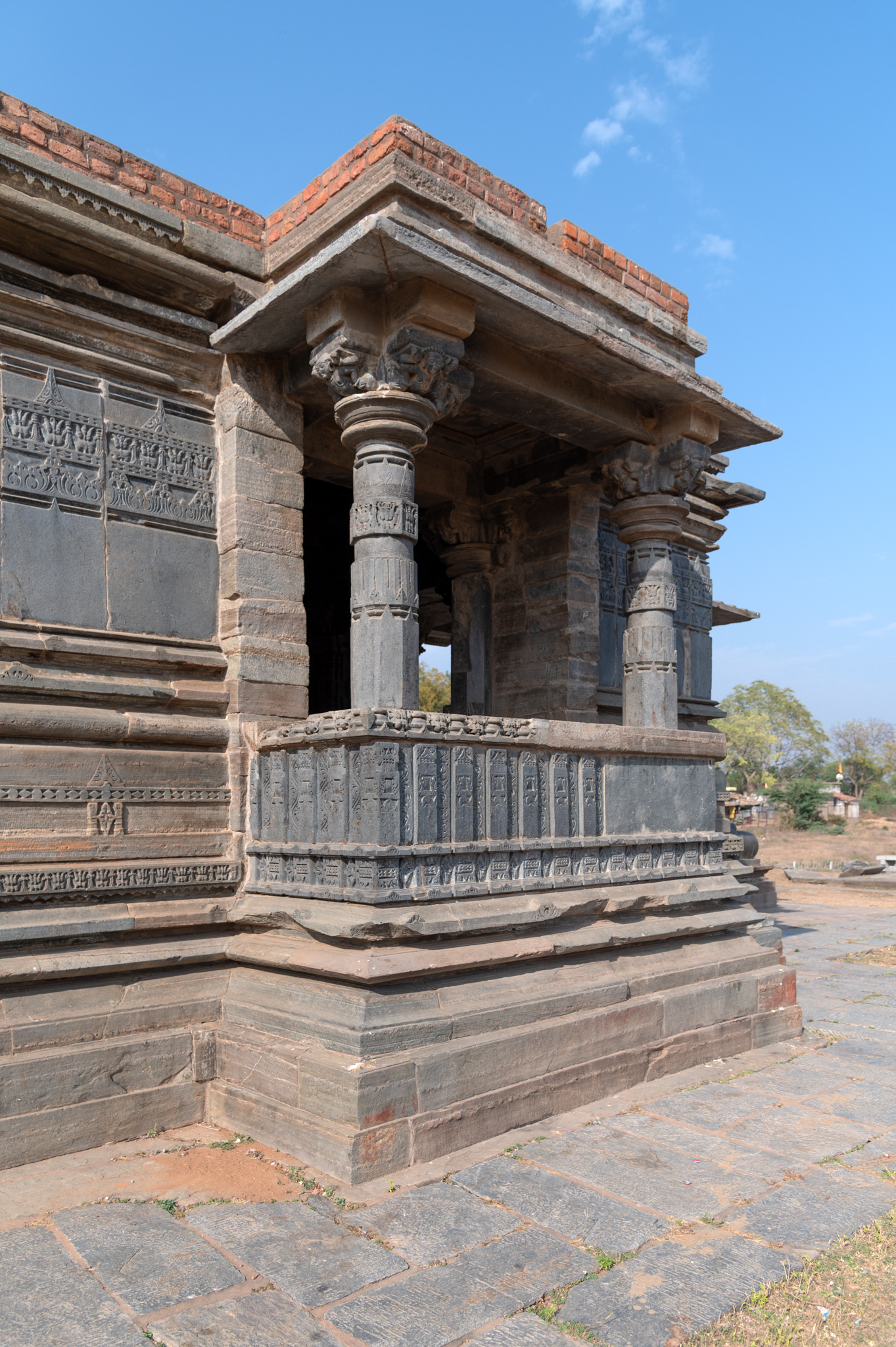 The Mandaleshwar Mahadev Temple's mandapa (pillared hall) is square in plan. It has lateral wings on its south and north sides. The exterior of the mandapa, the south-facing lateral wing, is shown here. This wing is a semi-open space. Its two front pillars support a protruding roof. The mandapa's exterior walls do not have any sculptures. At the jangha (wall) level, there are two decorative bands: the first is a foliage motif, and the second is a band of kirtimukhas (faces of glory).