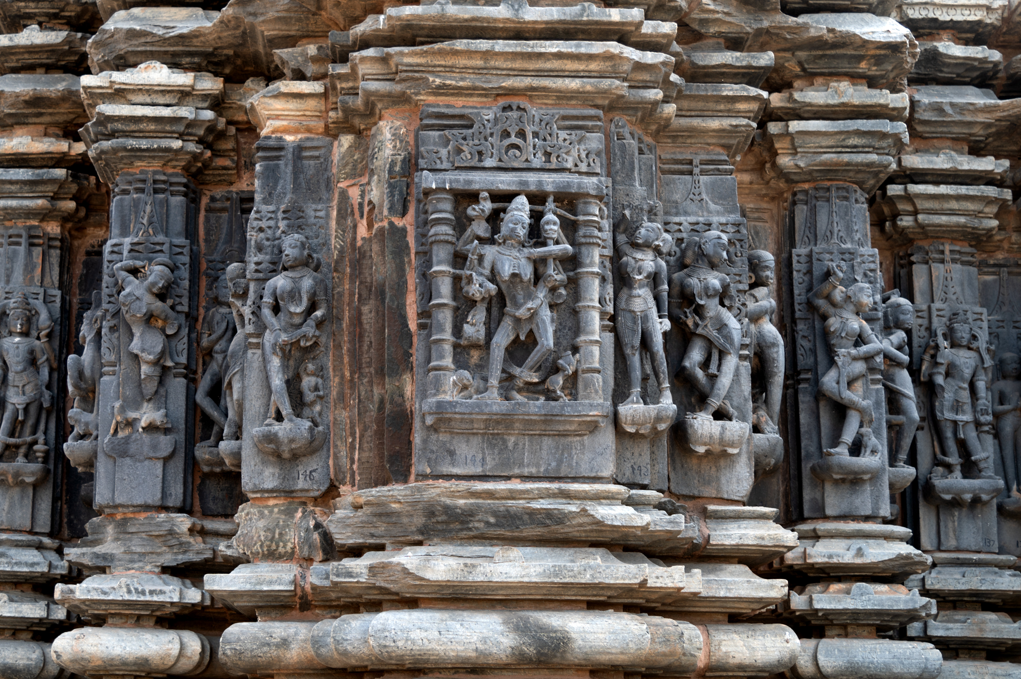 This is a closer view of the east jangha (wall) of the Kumbheshwar Mahadev Temple. The wall scheme is similar to the two north and south sides of the temple, with depictions of apsaras (celestial damsels), ascetics, and dancers on the intermediate and corner projections. The bhadra (central projection) features an image of Chamunda. Here, the depiction of Chamunda features a skeletal body, six hands, and a scorpion resting on her stomach. A supine human figure appears below the image. The goddess's other attributes, including a damaru (a small two-headed drum) and a khatvanga (a staff with a skull), are missing.