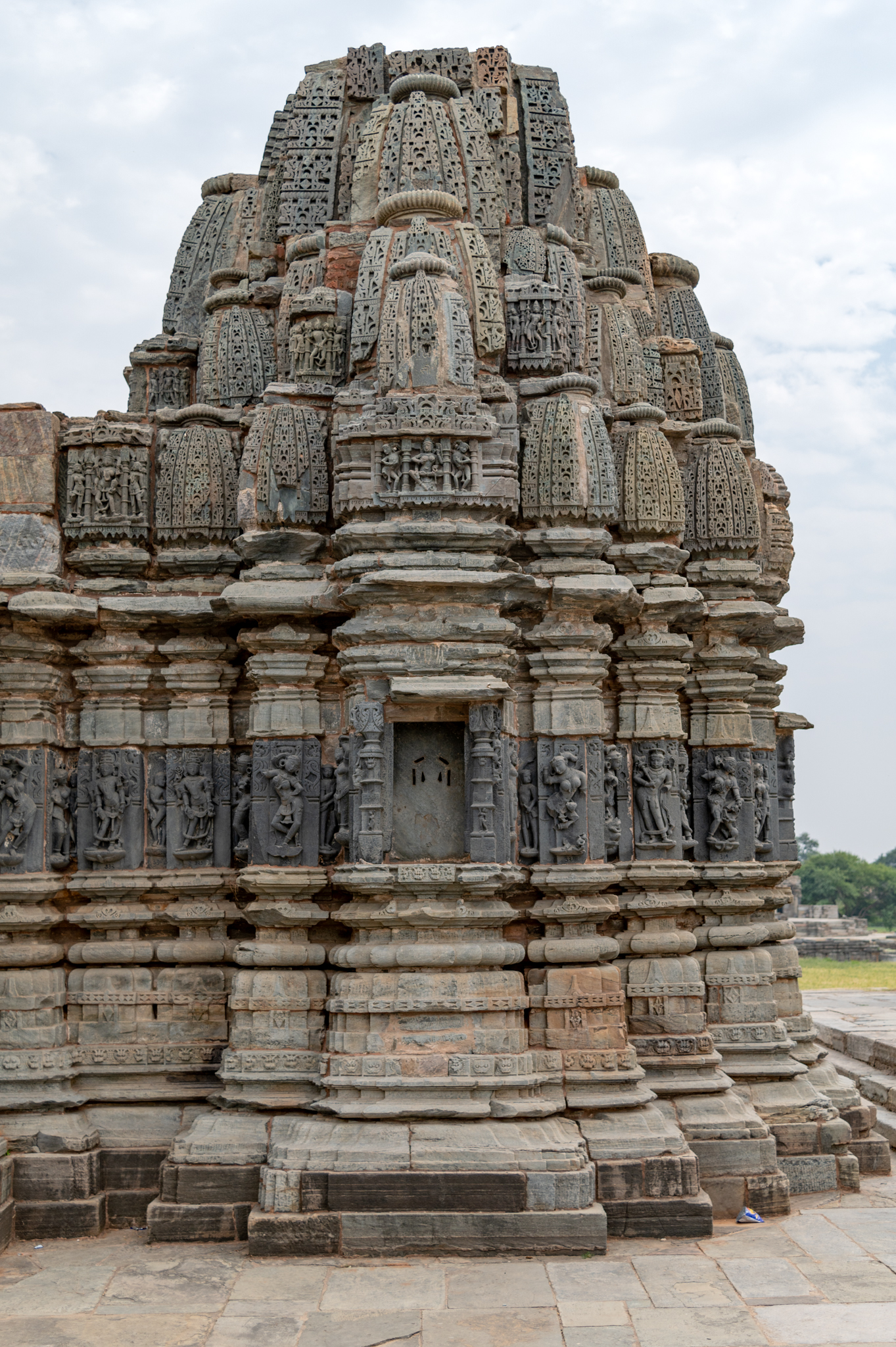 The Jain Temple's mulaprasada (main shrine) is visible here. The temple elevation has a vedibandha (basal mouldings) with minimal ornamentation, followed by a jangha (wall), which has figural sculptures and an intricately carved multi-spired shikhara (superstructure).