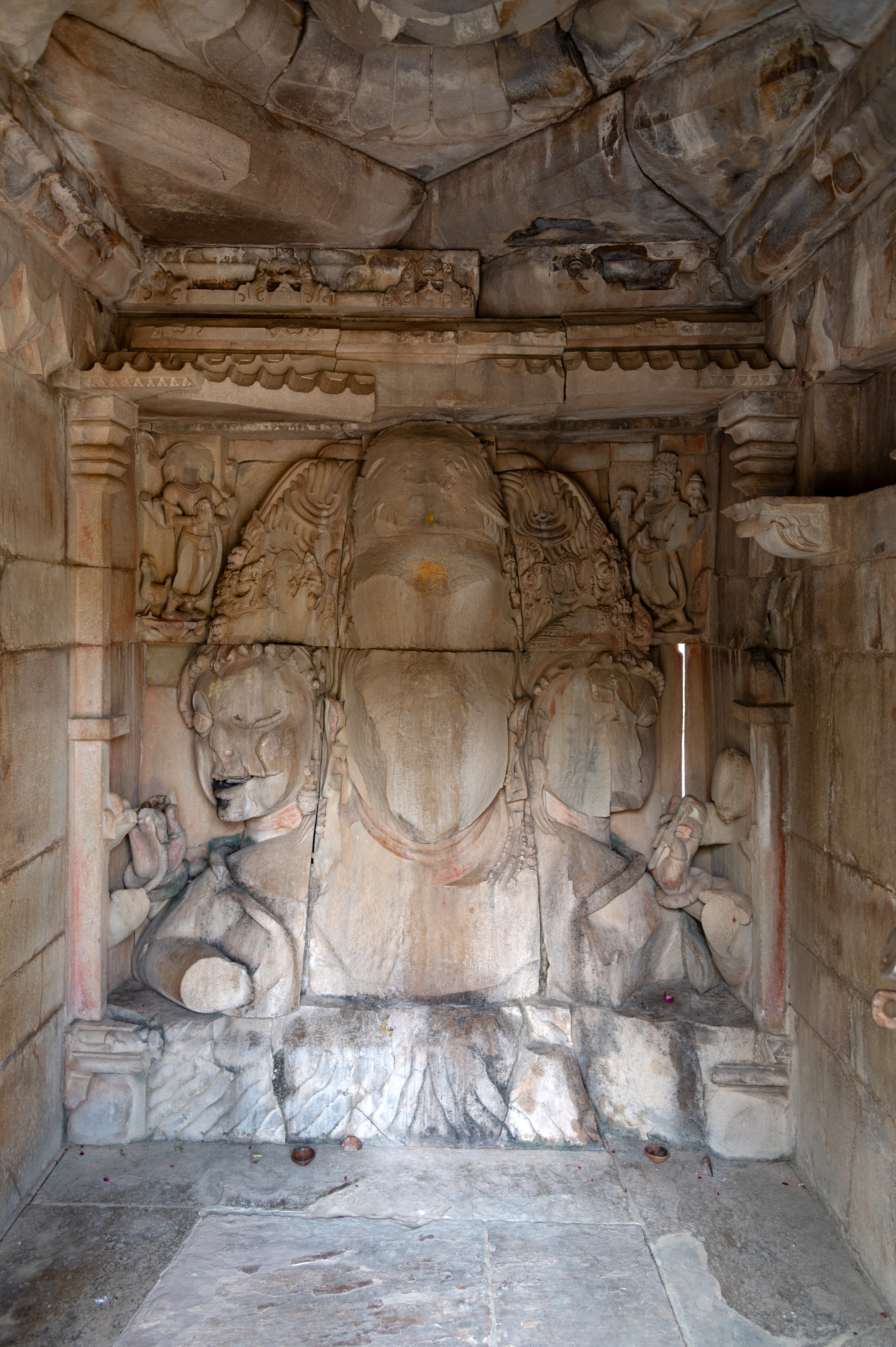 The garbhagriha (sanctum sanctorum) is a small chamber where a large bust of Sadashiva or Maheshamurti is installed. The bust depicts three faces. The frontal face is of Tatpurusha, which is completely rubbed off. On the proper right, Aghora is portrayed, representing the aggressive aspect, while the left face is Vamadeva, reflecting the feminine aspect. The Aghora face wears a jatamukuta (a crown of matted hair) adorned with skulls and snakes. He has raised, arched eyebrows, large bulging eyes, and a third eye on the forehead. The deity is depicted with fangs, but one can notice the smile on its face, and holding a snake in his hand. The right half of Vamadeva’s face is also chipped off. The deity is shown with a jatamukuta featuring kirtimukhas (face of glory) and rounded curls. The deity has bow-shaped eyebrows, beautiful eyes, and a third eye on the forehead.