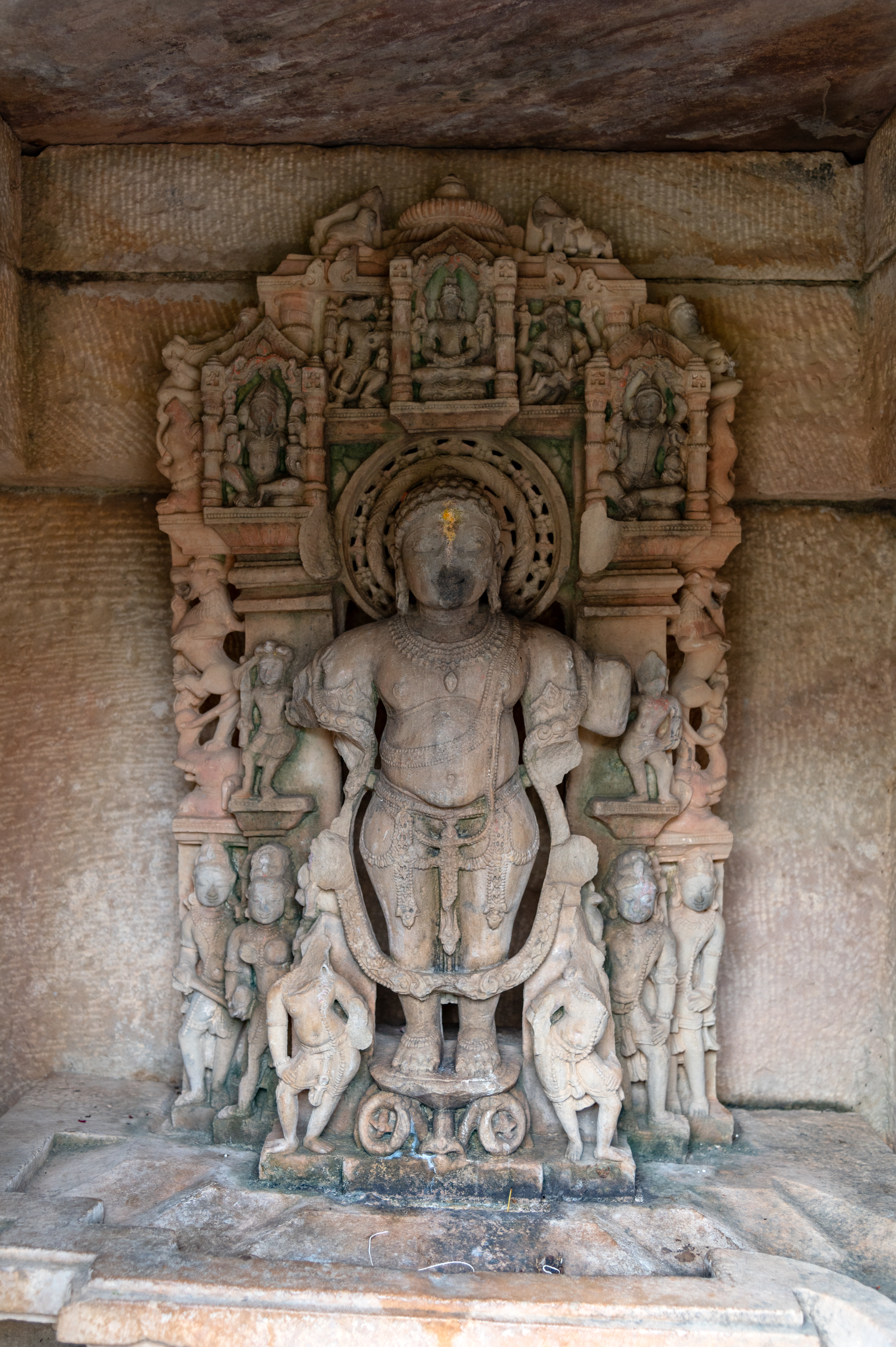 Vamana is depicted as a dwarf with a potbelly in the Vamana Temple. He had four arms which are all broken presently. He is heavily bejewelled and wears a long vanamala (forest flowers garland). He is accompanied by six attendants who can be seen at the bottom. There are two more figures on his sides, near his arms. Notably, miniature niches at the top of the sculpture house representations of the Hindu trinity; Brahma, Vishnu, and Mahesha.