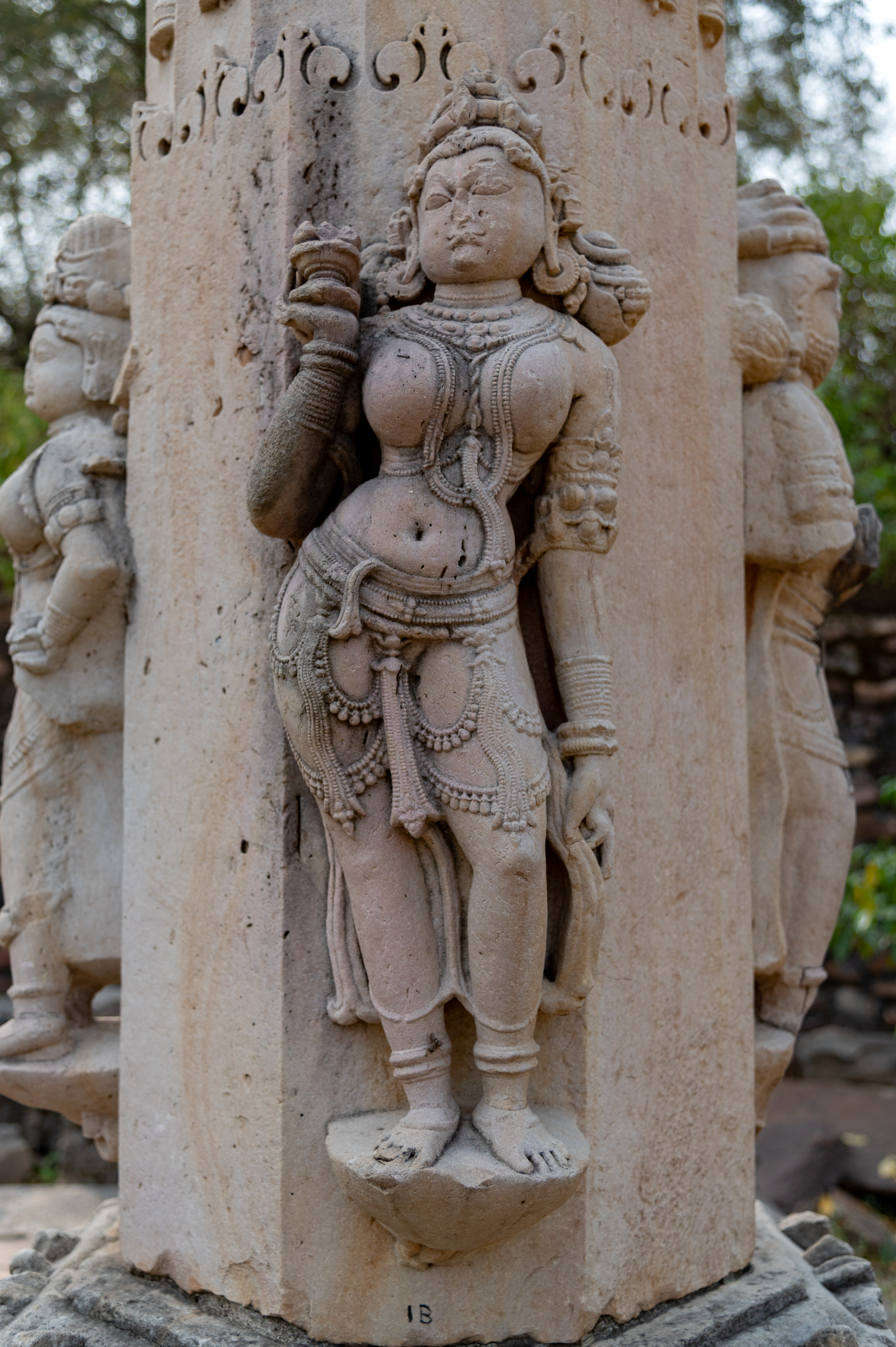 The shaft of the torana (ornamental gateway) displays four figures on each of its four sides of the octagonal base, while the remaining four sides are left plain. Each side of the shaft features three female figures and one male figure. One of the female figures is shown standing, holding a bowl and from her other hand, she gracefully holds her drapery. The figure embodies feminine beauty, characterized by her curvaceous figure, elegant hairdo, and jewellery.