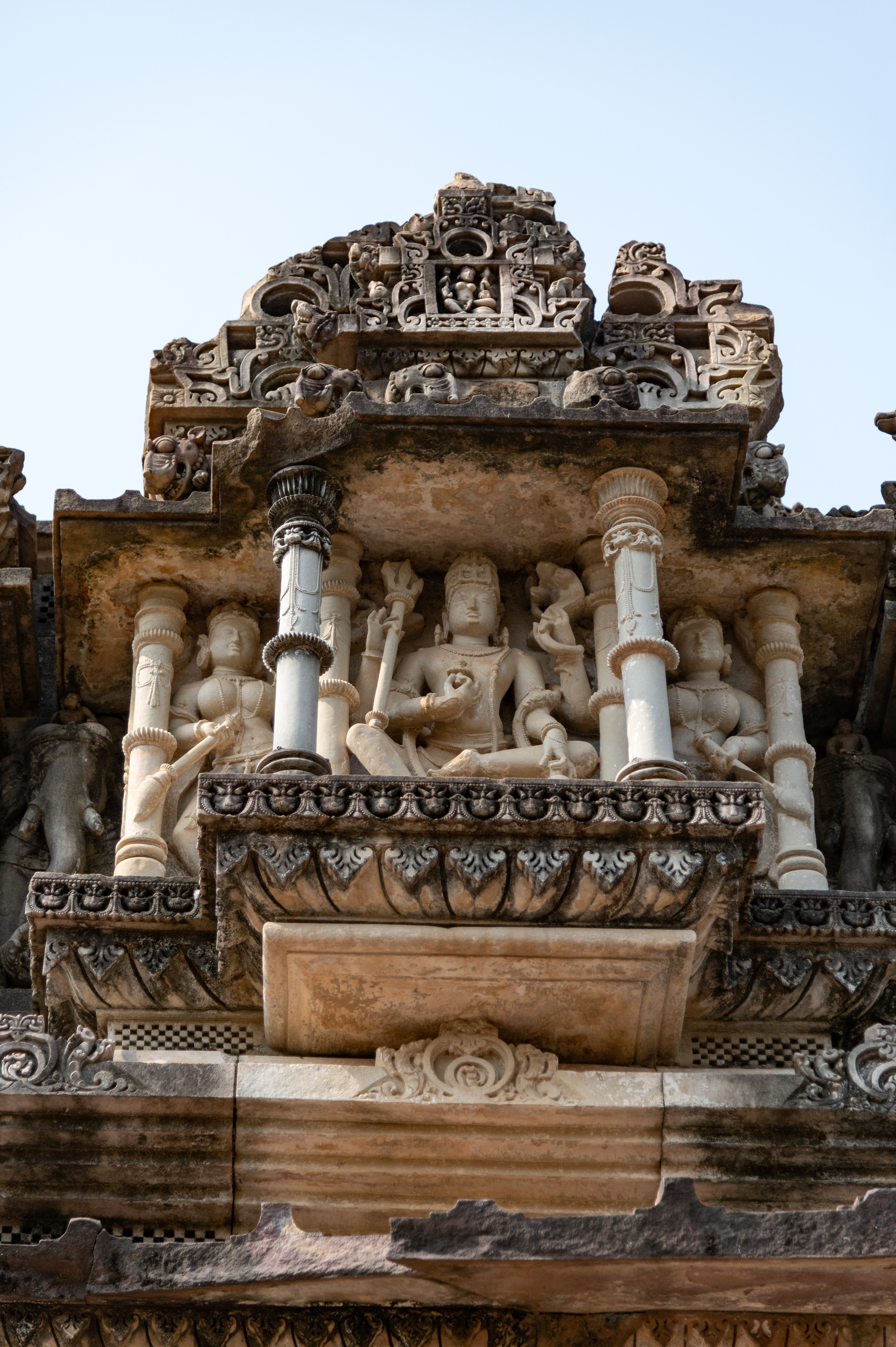 The eastern pediment of the mukhamandapa’s (front porch) shikhara (superstructure) depicts a beautiful, seated figure of Shiva. The four-armed Shiva holds a trishula (trident) and a sarpa (snake) in his upper hands. In his lower left hand, he holds a kamandalu (water pot) and a pushpa (flower) in his lower right hand, which is placed near his chest. Shiva is flanked by two female fly whisk bearers.