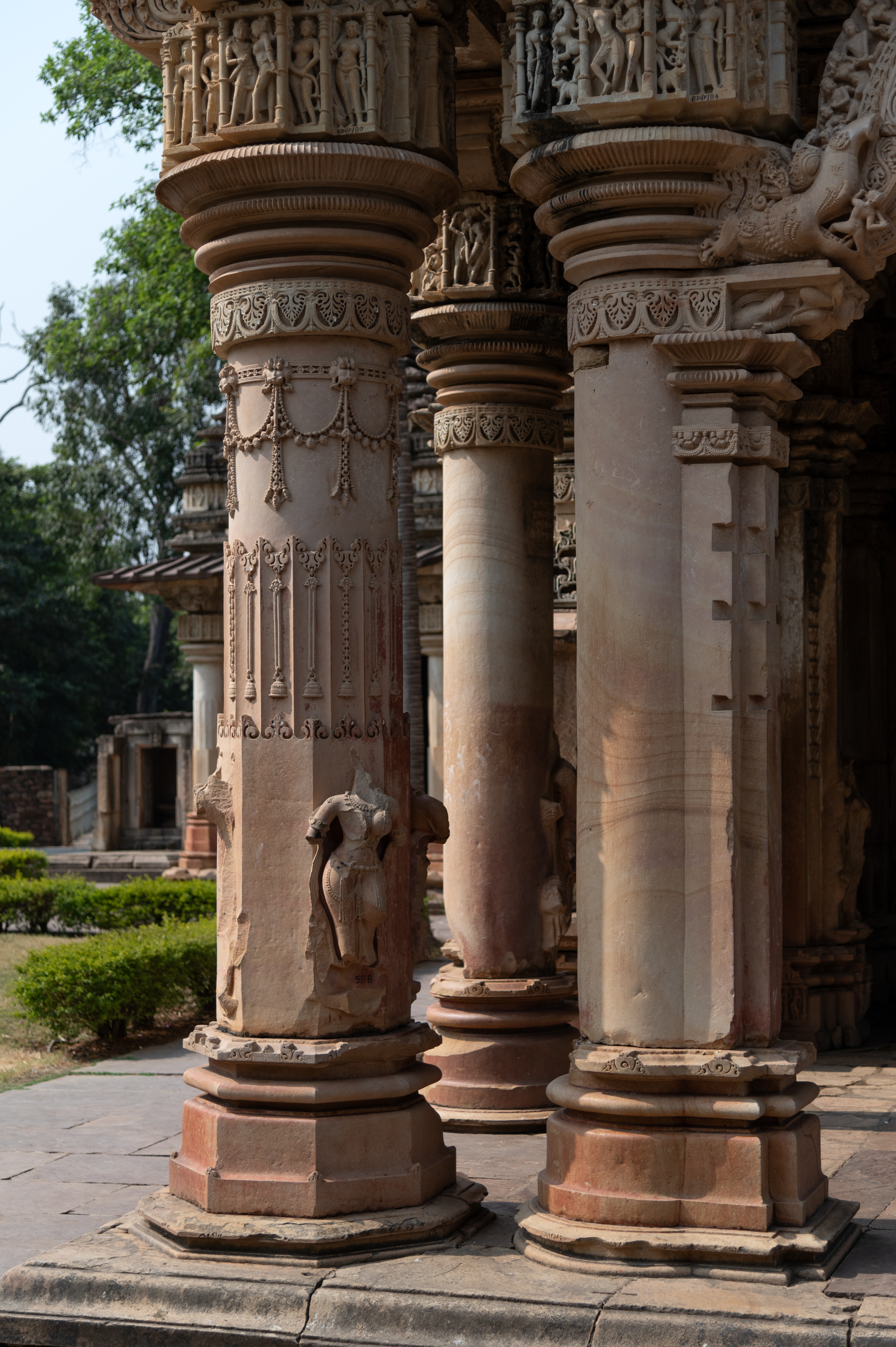 The shafts of the front pillars are adorned with loops of bells and hanging bells. The pillars are adorned with the graceful standing figures of sursundaris (celestial maidens) which are currently broken and damaged. The figural panel at the top of the cushion abacus depicts sursundaris, dancers, and couples.