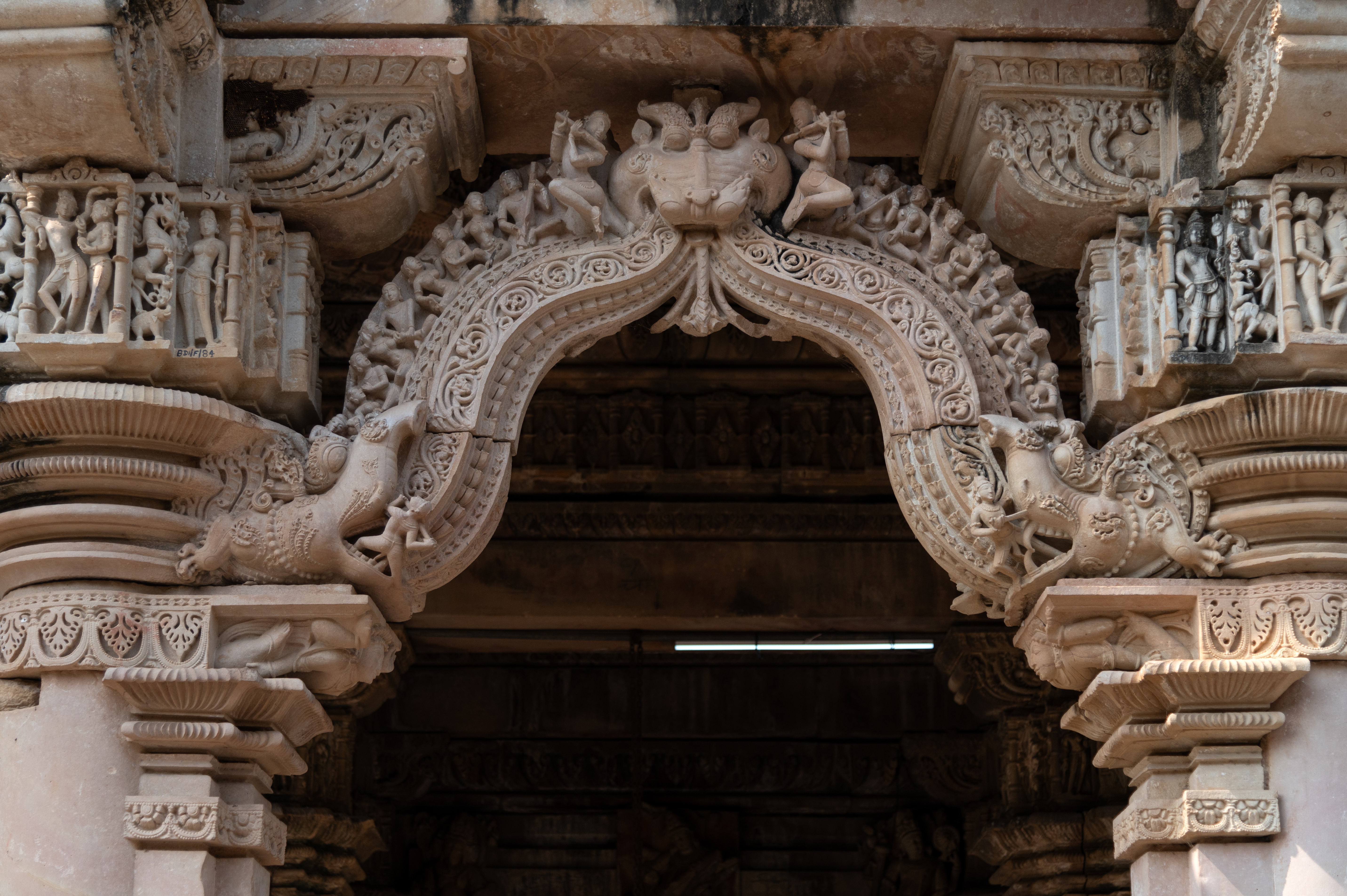 The mukhamandapa (front porch) of the Ghateshwar Temple has an entry through an intricately carved makara-torana (ornamental arch adorned with intricate carvings of mythical crocodile creatures). The torana (ornamental arch) shows makara (crocodile) on each end. These makaras are being attacked by knights with a sword in their mouth. The floral bands emitting from the mouths of the makaras form a pointed arch, above which musicians are shown seated, playing musical instruments, and singing. At the apex, a kirtimukha (face of glory) flanked by two flute players is depicted.