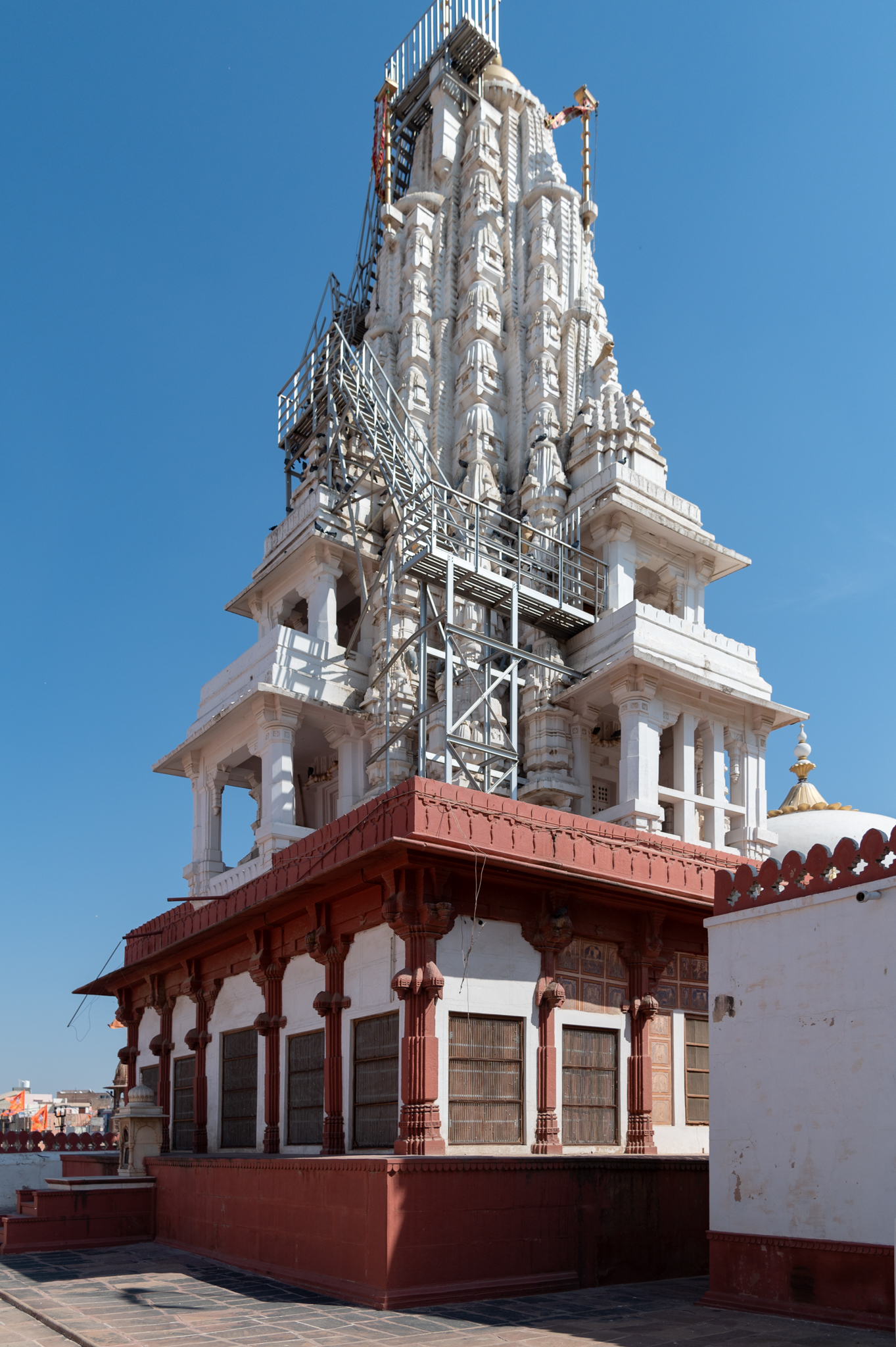 View of the entire elevation of the Bhandasar Temple from the southwest corner of the complex. The temple proper is painted in bi-chrome, red ochre and white, but the shikhara (superstructure) of the temple is only in white. The plinth of the temple looks like it was built in brick. However, the entire structural core is built in stone. The base of the shikhara has balconies on two terrace levels of the temple. These can be accessed from inside the temple. It is said that one can see the entire Bikaner city from these balconies.