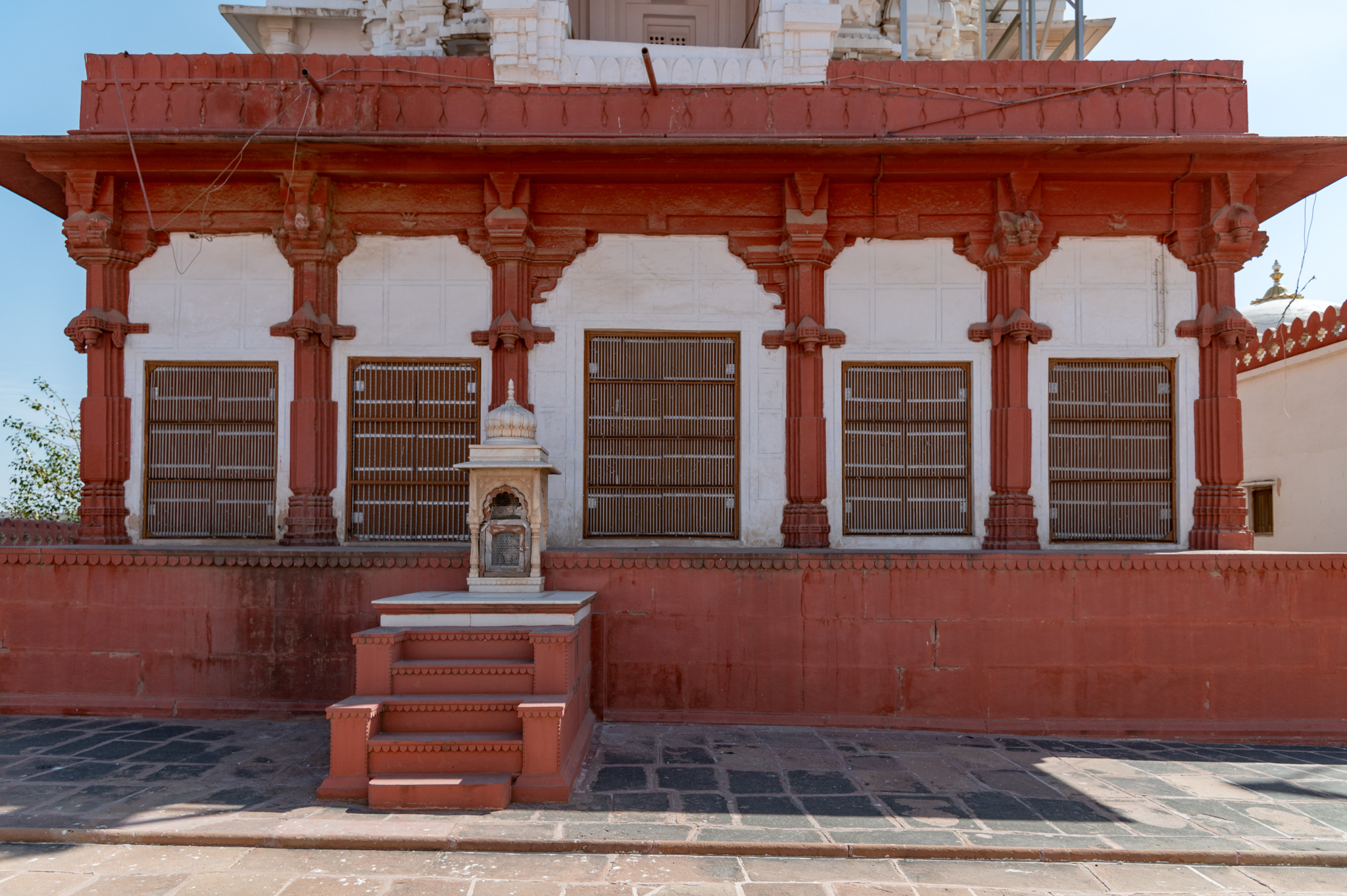 Seen here is the west-facing elevation of the Bhandasar Temple. The exterior walls of the temple are divided into five bays, of which the central bay is wider than the two bays flanking it on either side. The doorways of these walls, when open, make the interiors of the temple semi-open and light up the interiors. The blind foliated arches of each bay have rectangular patterns that might have had paintings at some point, but at present, only the outlines of these rectangles survive. At the centre of the plinth, there is a small chhatri (memorial), which was probably added to the temple later.