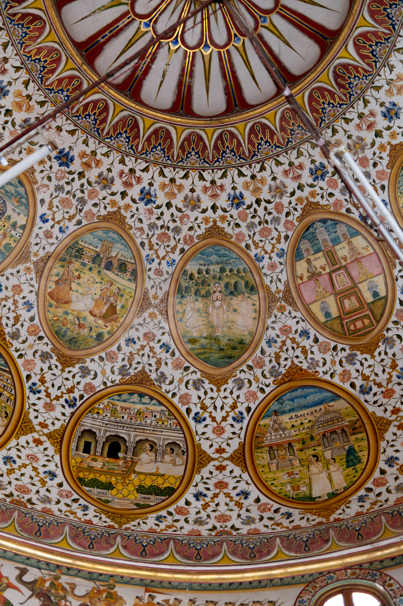 Seen here are five paintings in the oval cartouches in the ceiling of the mandapa (pillared hall) of the Bhandasar Temple. The oval shapes of the paintings are designed like mirror frames. This style is influenced by European paintings, especially the Company style paintings produced during the 18th – 19th centuries CE. The lower row depicts stories of Jinadatta Suri’s miracles when he made a dead cow, which was in front of the Jain temple, alive and put it in front of a Hindu temple. The left oval has a painting of Jinadatta Suri miraculously saving the Mughal prince. In the upper row are paintings of (left to right of the viewer) the sacred Meru mountain, Bahubali and Rishabnatha.