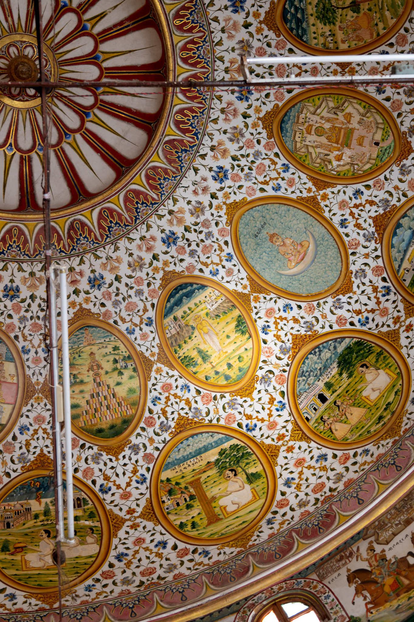 A closer view of the paintings in the oval cartouches in the ceiling of the mandapa (pillared hall) of the Bhandasar Temple. Seen here are many ovals depicting different stories. The lower set has stories of miracles of Jinadatta Suri, like saving a child’s life, procuring the sacred scriptures hidden inside a pillar, and capturing the lightning in his alms bowl. The upper circle has Jain monks and auspicious symbols for Jain tradition, like Chaude Rajlok and Mokhs Parinirvana, enunciating the worldly life. The oval shapes of the paintings are designed like mirror frames. This style is influenced by European paintings, especially the Company style paintings produced during the 18th – 19th centuries CE.