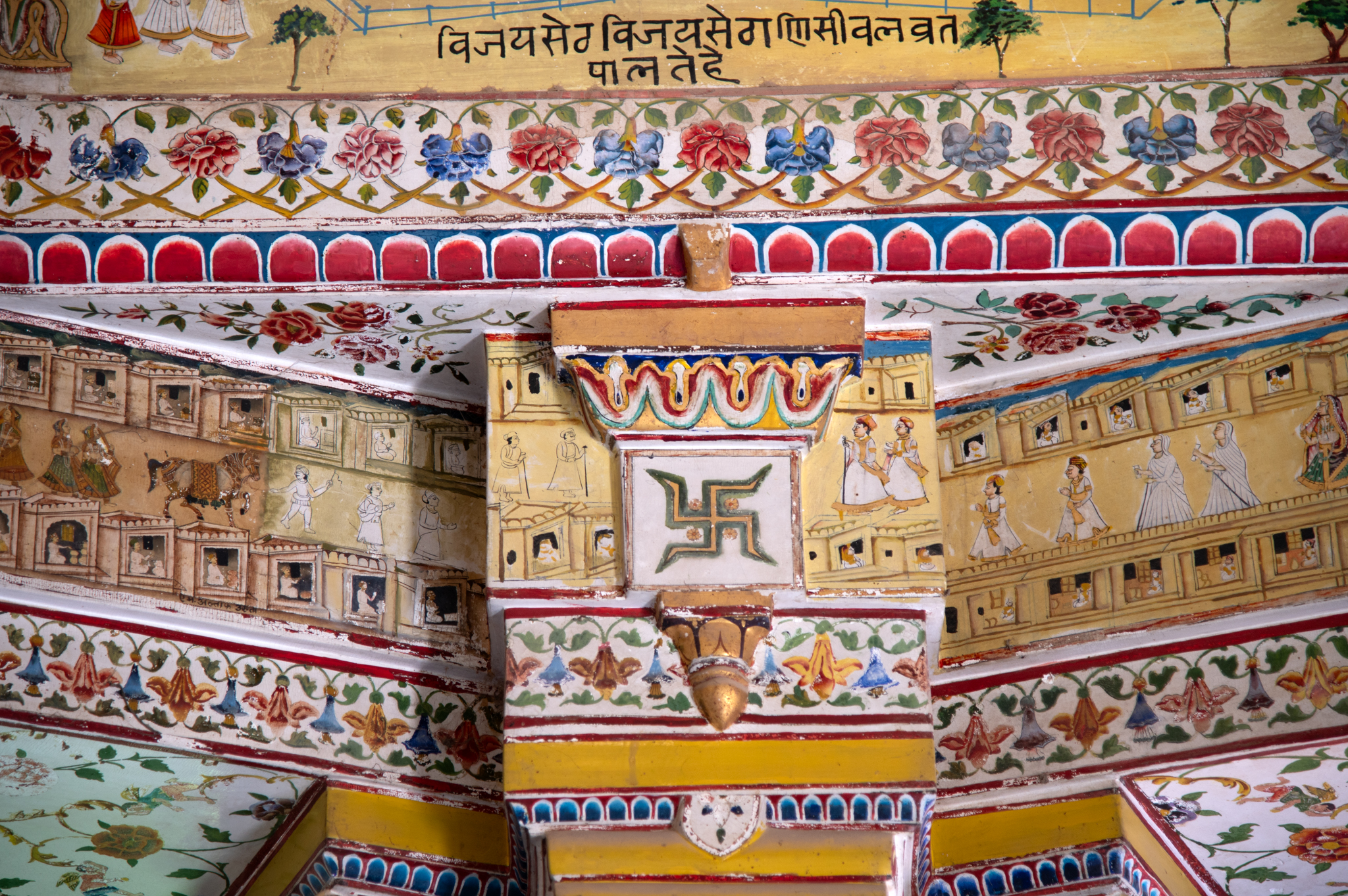 The pillar capital in the picture has a Swastika symbol, one of the eight auspicious symbols of Jainism. It is surrounded by floral motif bands. The band flanking the Capital has a depiction of scenes from the city of Bikaner where people are seen going to the market, travelling on elephant back, shops in marketplaces, etc.