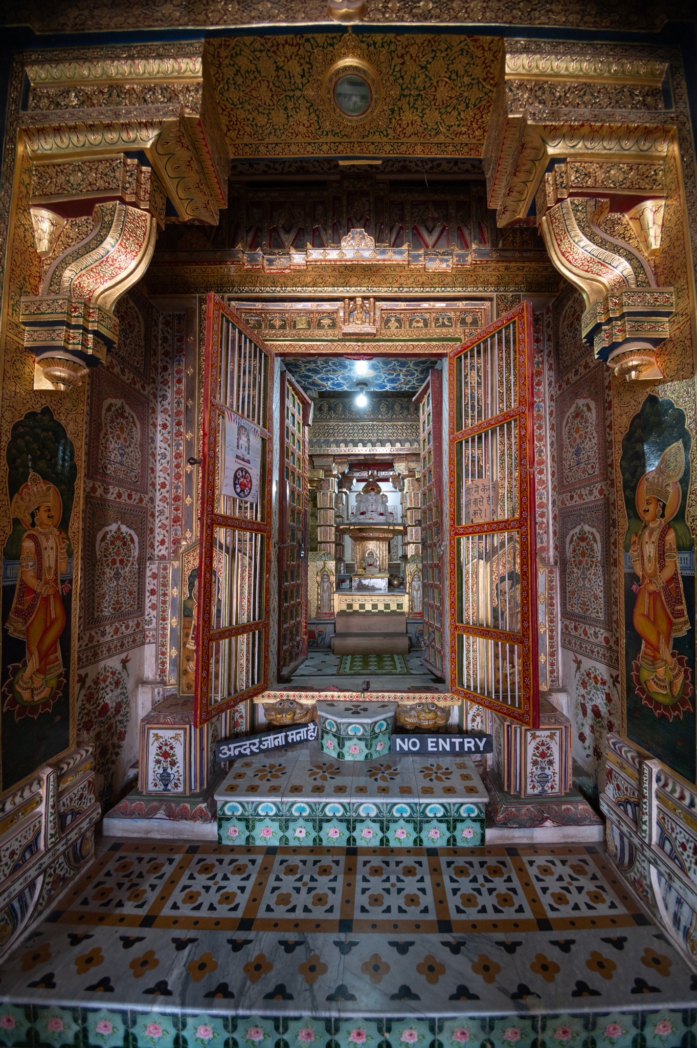 Seen here is the principal east-facing entrance to the garbhagriha (sanctum sanctorum) of the Bhandasar Temple. The original fabric of this entrance has been enveloped in golden embossing. The pilasters, doorway and dvarashakha (architrave) are adorned with golden meenakari (enamel) work. Intricate details of the floral motifs cover the entire surface of the entrance. The wooden doorway has attendants painting on it.