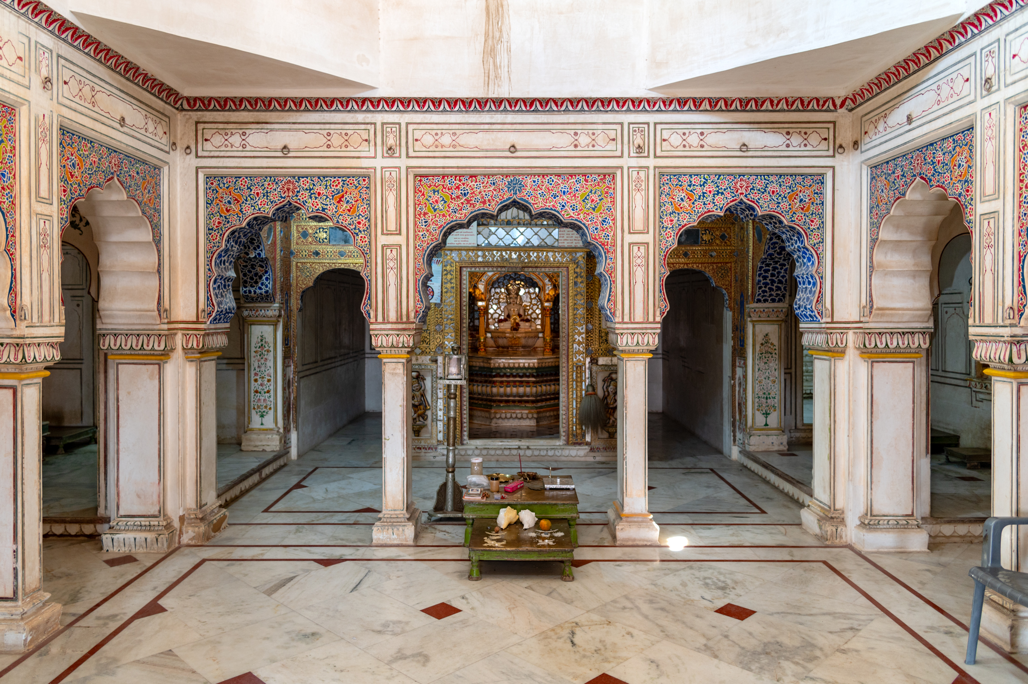 This is the mandapa (pillared hall) of the Adinatha Temple, which has a quadrangle at the centre surrounded by rectangular archways on its northern and southern sides. To the west, the mandapa is connected to the garbhagriha (sanctum sanctorum) via an antarala (antechamber). The tripartite division of the arches is the same on all sides. The arches are supported by piers on the corners of the row. The base colour of the interiors of the mandapa is white, on which it has beautiful paintings done using multiple colours.