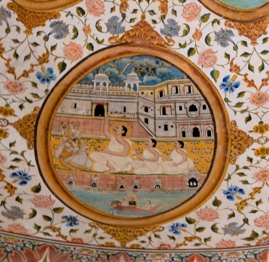 The fourth oval painting (in clockwise order) in the lower/outer circle in the ceiling of the mandapa (pillared hall) is an episode about Jinachandra Suri’s life. The inscription on the viewer’s left side of the painting is lost, hence the exact episode is unknown. The painting depicts a Mughal official with his attendants bowing down to Jinachandra Suri, accompanied by his disciples, during a religious gathering. The acharya (those who impart knowledge) is seen pointing towards the sky, where lightning strikes. The episode is happening in front of a haveli. In the foreground of the painting, two Mughal officials, possibly sitting on horses, are depicted. A similar episode is found in the life of Jinadatta Suri, where he saves his followers from drowning in another part of the world, showcasing his knowledge and power. The remnant inscription reads, ‘Shri Jinachandra Suri ji.’