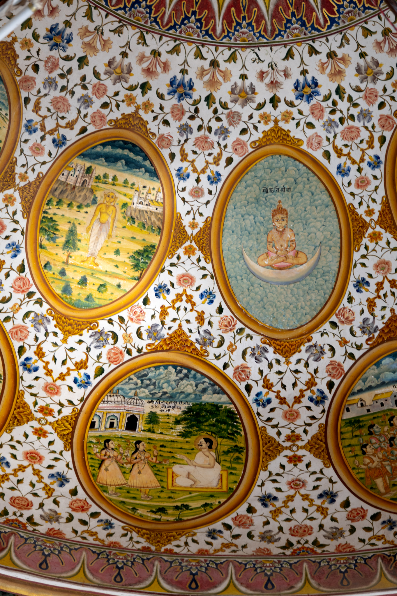 One of the most well-known stories of the miracles of Jinadatta Suri, the eleventh oval painting (in clockwise order) in the lower/outer circle in the ceiling of the mandapa (pillared hall) depicts this episode of the story of sixty-four yoginis. The story takes place in Ujjain. Once, Jinadatta Suri was amidst a religious discourse when he foresaw that sixty-four yoginis were coming his way to disturb the gathering. The yoginis arrived disguised as laywomen. Jinadatta Suri asked his disciples to spread sixty-four mats for the yoginis, and using his powers, the acharya (those who impart knowledge) glued the yoginis to the mat. This way, the yoginis were compelled to attend the religious discourse and eventually became disciples of Jinadatta Suri. According to Lawrence Babb, this episode depicts the Jain attempt to tame the tantric goddesses. The inscription reads, ‘Shri Jinadatta Suriji 64 joganiya saji.’