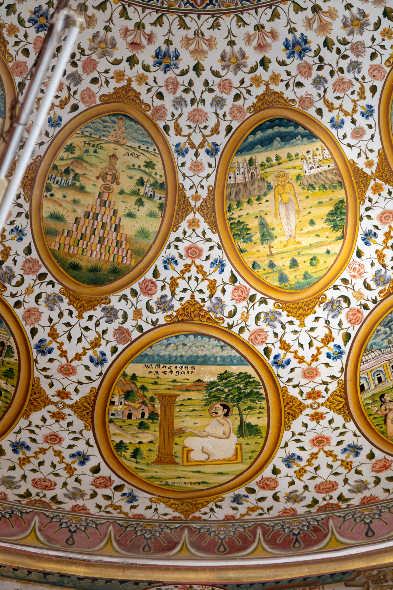 The twelfth oval painting (in clockwise order) in the lower/outer circle in the ceiling of the mandapa (pillared hall) is the episode of Jinadatta Suri when he procured texts which were hidden in the pillar by a Jain ascetic several years ago. No one knew the location of the sacred scriptures, but the acharya (those who impart knowledge) could discern it using his magical powers. The pillar containing the sacred scriptures is depicted as a fluted column. This painting bears the name of the painter, M. Altaf Usta. The word ‘Usta’ used as a suffix here comes from the word ustad, meaning expert.