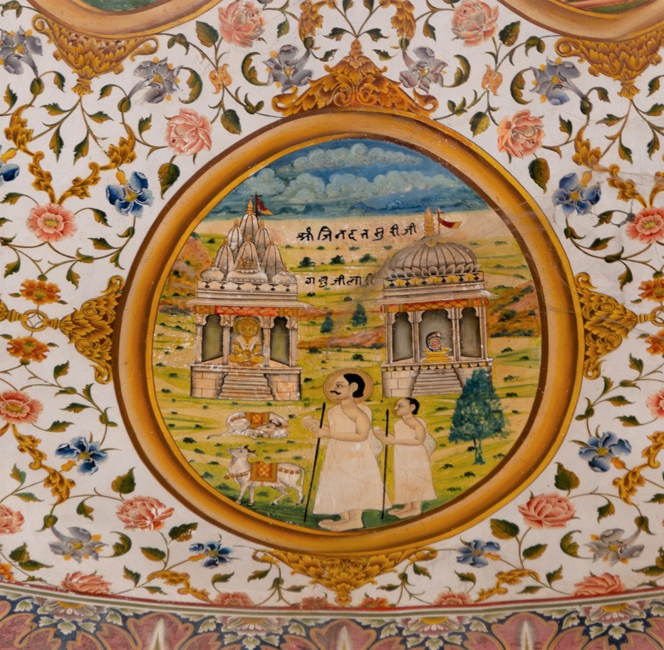 The fourteenth oval painting (in clockwise order) in the lower/outer circle in the ceiling of the mandapa (pillared hall) is another story about the miracle of Jinadatta Suri. Once, somewhere in Gujarat, with the evil intent of troubling the Jains, some people placed a cow in front of the Jain Temple. Jinadatta Suri, using his powers, placed the cow in front of the Shiva Temple. Interestingly, the Hindu temple, with a Shiva linga inside, is shown with a domical spire, while the Jain Temple is shown with a Tirthankara enshrined in it, featuring a multi-spired roof. The painting includes two cows, one sitting in front of the Jain Temple, and the other standing in front of Jinadatta Suri. Jinadatta presumably also restored life to the cows. A disciple accompanies him. The inscription reads, ‘Shri Jinadatta Suri ji Gavu ji la hi.’