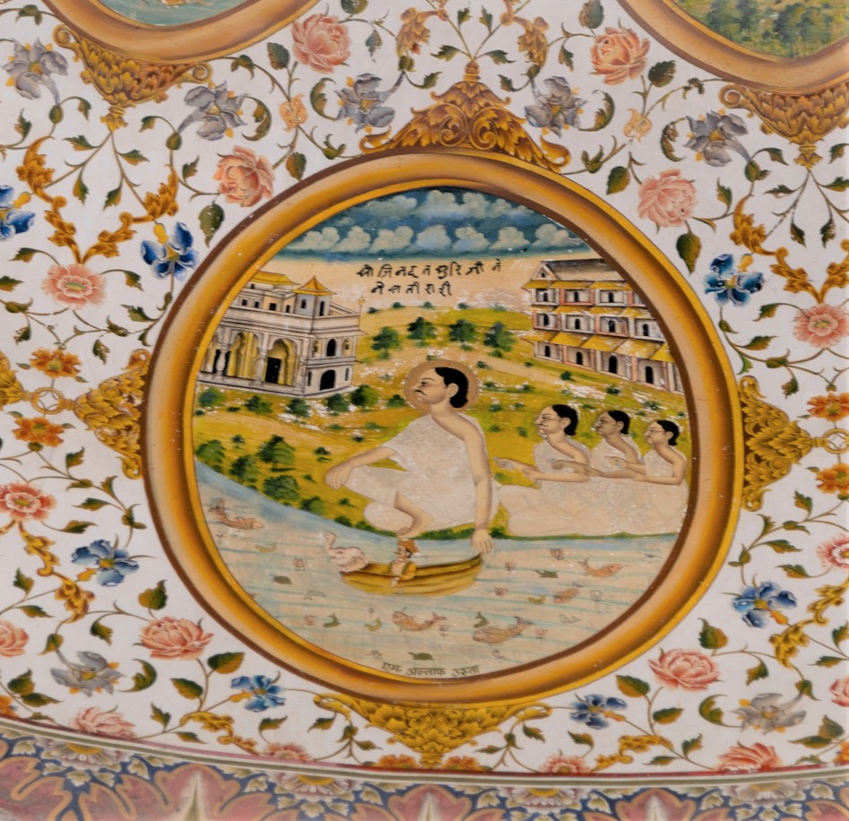 As per the inscription on the fifteenth oval painting (in clockwise order) in the lower/outer circle in the ceiling of the mandapa (pillared hall), it depicts Jinadatta Suri reviving a Mughal prince. The story, commonly told in Jain traditions, recounts that once a Mughal prince fell from a horse and died. Jinadatta Suri commanded Vyantar Dev (forest celestial beings) to enter the dead prince’s body and bring him back to life. The Mughal prince is shown sleeping on a charpahi (four-legged bed) in the courtyard of the haveli (mansion). In the background, several buildings are depicted, suggesting a city settlement. Some of these buildings exhibit European architectural designs. The inscription reads, ‘Shri Jinadatta Suri Ji mugal putar ji la yo.’