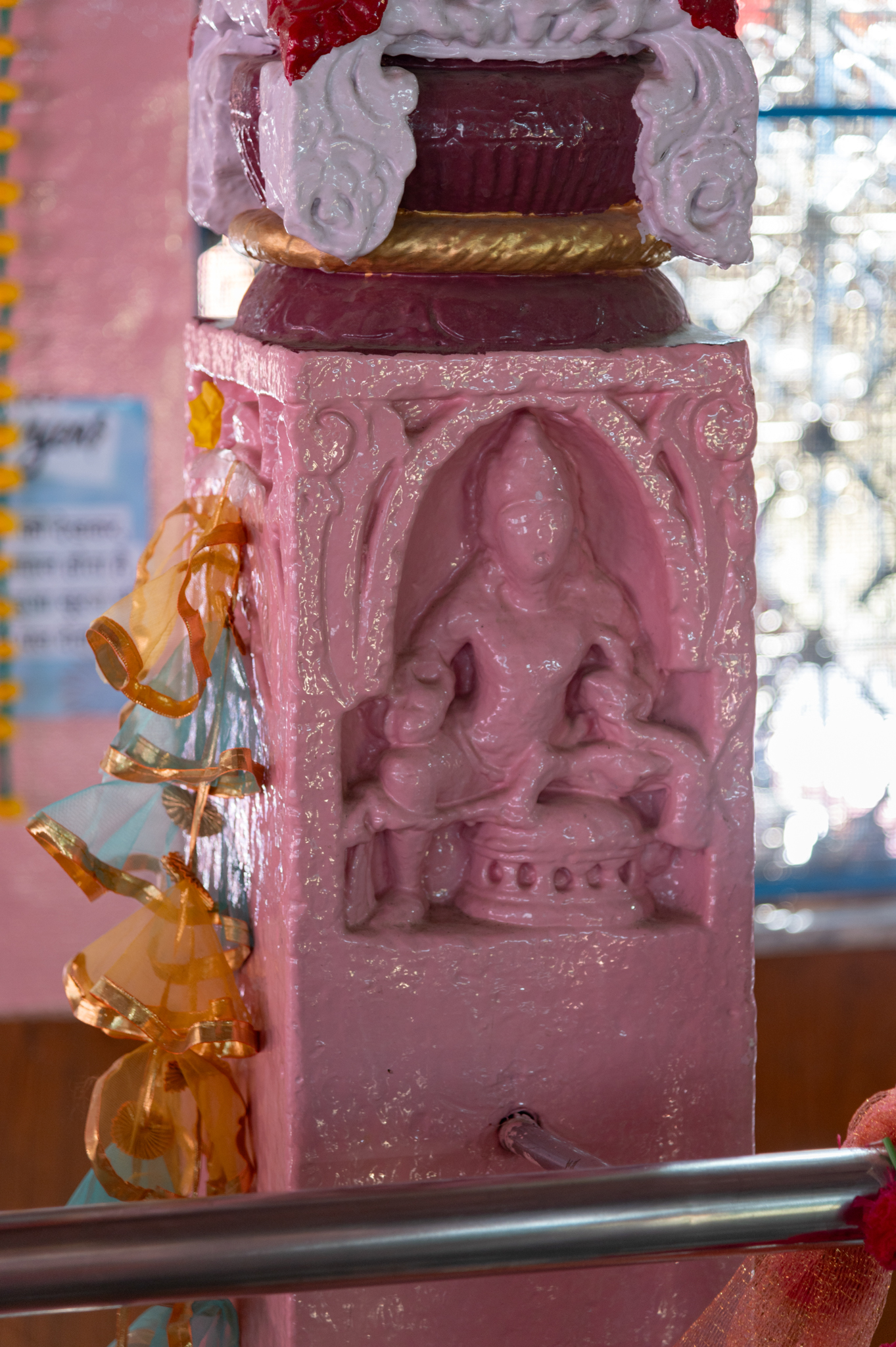 Seen here is a sculpture of a female on the south-facing (inner) side of the pillar in the northwest corner of the mandapa (pillared hall) of the Suswani Mata Temple. It is said that there used to be an inscription below this sculpture, which is probably not visible now because of the later added paint. This inscription was supposed to be the sole epigraphical evidence dating the construction of the temple in the 10th century CE. A sculpture carved in deep relief (probably a female) is shown seated on a pedestal. The sculpture is two-handed only and is shown wearing a mukuta (crown).