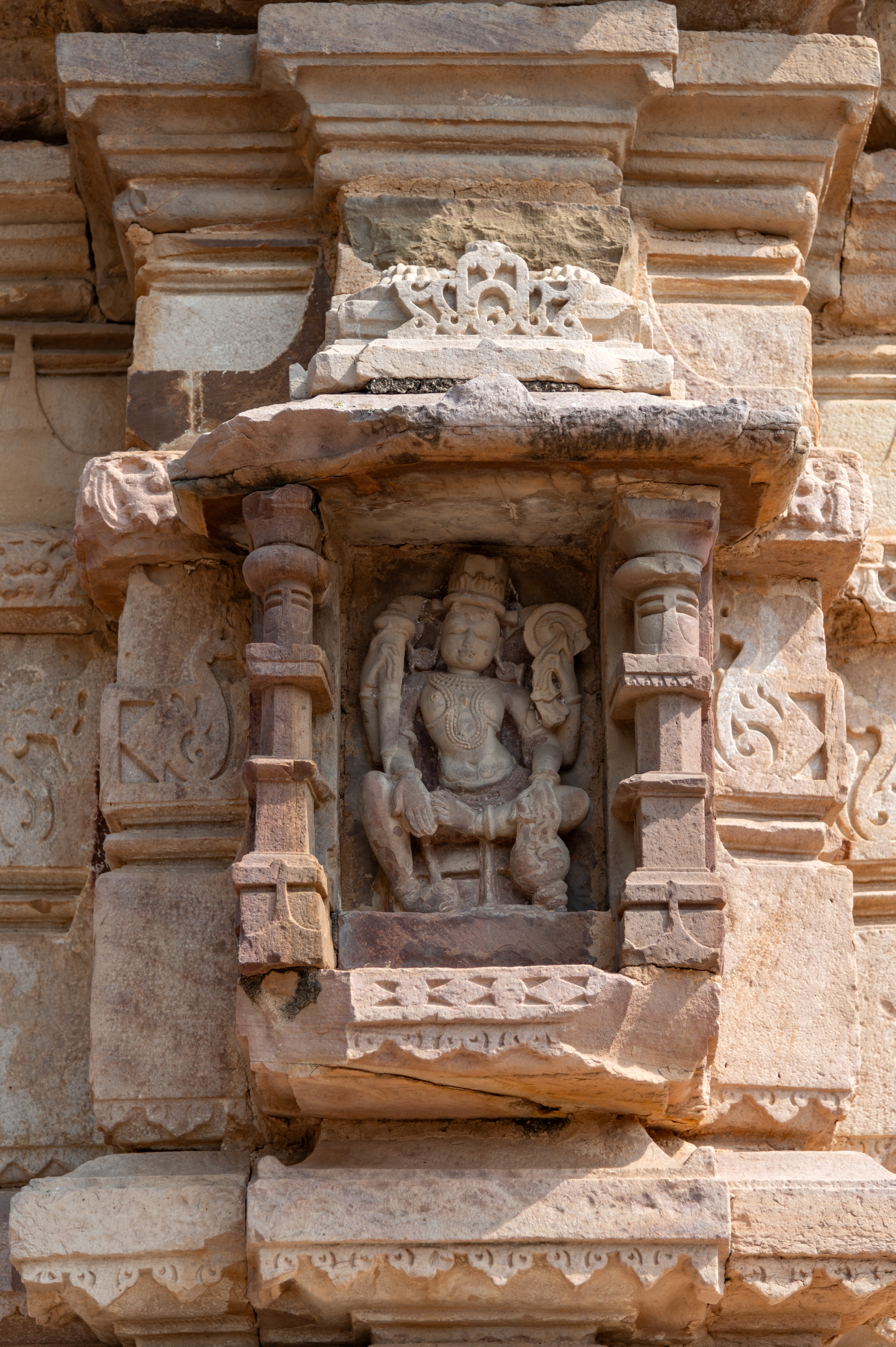 On the western bhadra (central offset) of the garbhagriha (sanctum sanctorum) of the Hazareshwar Temple, there is a depiction of Vishnu seated on a pedestal. Vishnu is portrayed with four arms, holding a gada (mace) and a chakra (discus) in his upper hands, and a shankha (conch shell) in his lower left hand, while his lower right hand is in the Varada hasta (hand gesture of conferring a boon) posture. He is adorned with elaborate accessories, including a kiritamukuta (conical cylinder-shaped crown generally attributed to Vishnu iconography), large earrings, a broad necklace, a chest band, a heavy girdle, bangles and anklets enhancing his divine appearance.