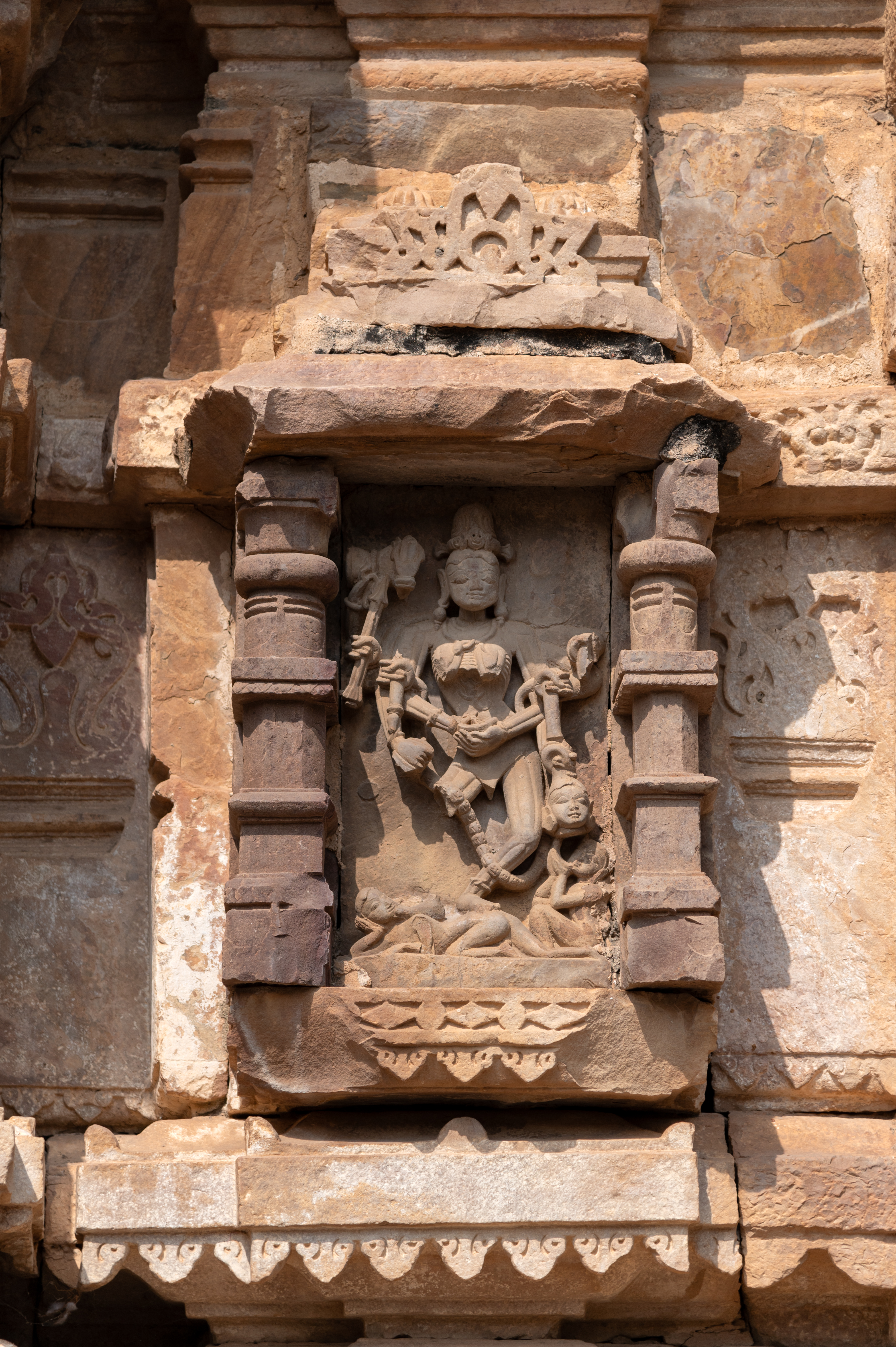 A depiction of the goddess Chamunda is there on the southern wall of the antarala (vestibule or antechamber) of the Hazareshwar Temple. Chamunda, portrayed with twelve arms, stands upon a human body. Skeletal depiction with desiccated breasts and a sunken belly containing a scorpion. She wears a triangular loin cloth to cover her lower body. She has a jatamukuta (crown of matted hair) adorned with a skull at the front. Large earrings dangle from her elongated earlobes, and a snake wraps around her neck and ankles. In her right hand, she holds a trishula (trident), damru (double-sided drum), arrow, a broken weapon with a staff and a bell (ghanta). In her left hand, she carries a snake and a broken weapon with a staff. Her left frontal hand holds a bowl, while her right frontal hand touches it. At the bottom left, a human figure is depicted seated, drinking blood dripping from a severed head into a bowl.