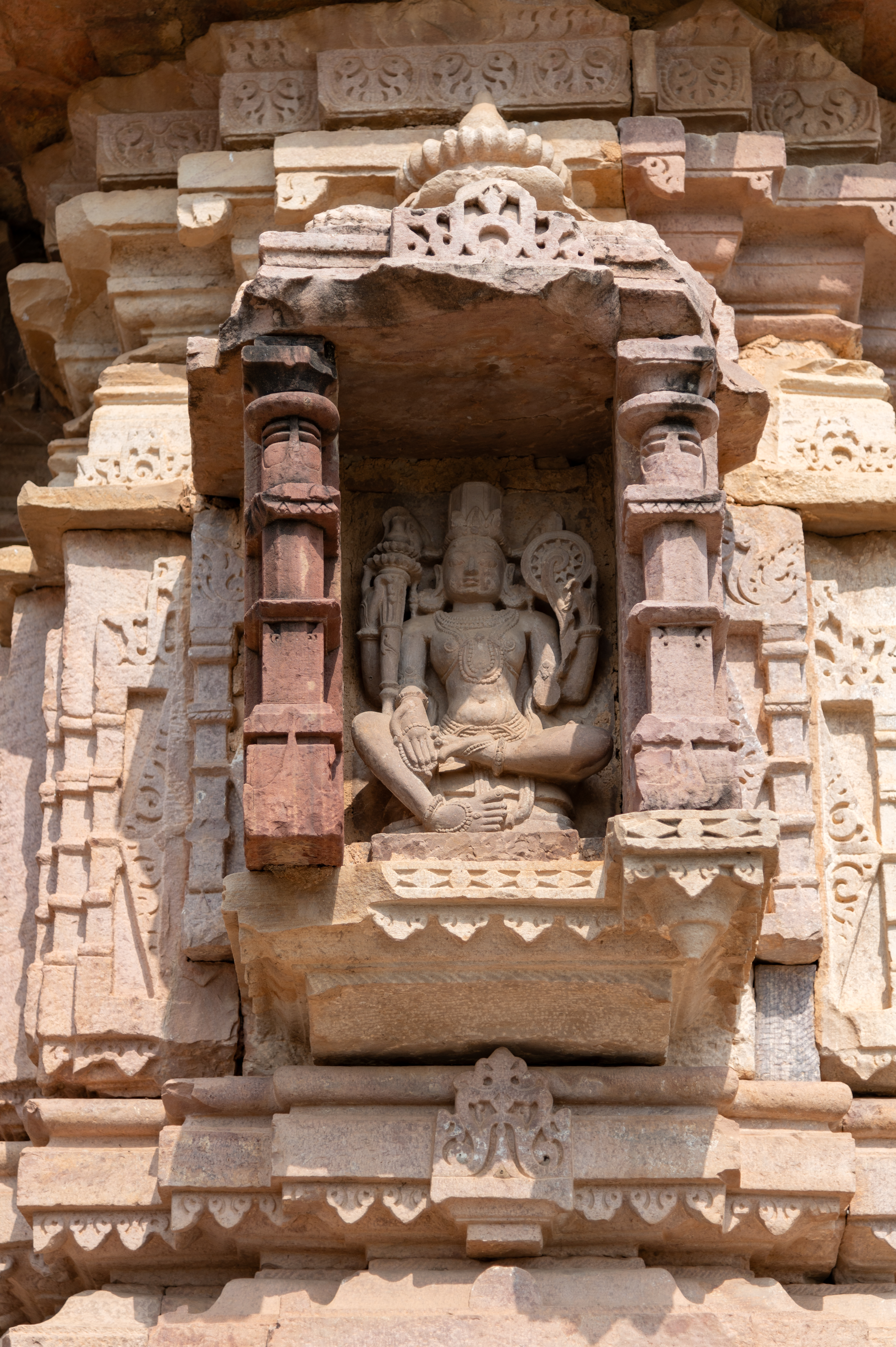 One of the bhadras (central offsets) of the garbhagriha (sanctum sanctorum) of the Undeshwar Temple features a seated Vishnu depicted with four arms. Adorned with a crown, Vishnu holds a gada (mace) and a chakra (discus) in his upper hands. In his lower right hand, he bears an akshamala (rosary) while also displaying the Varada hasta (hand gesture of conferring a boon). Unfortunately, his lower left hand is damaged. The figure is adorned with elaborate jewellery, including large circular earrings, a broad beaded necklace, a breast band, a girdle, bangles and anklets.