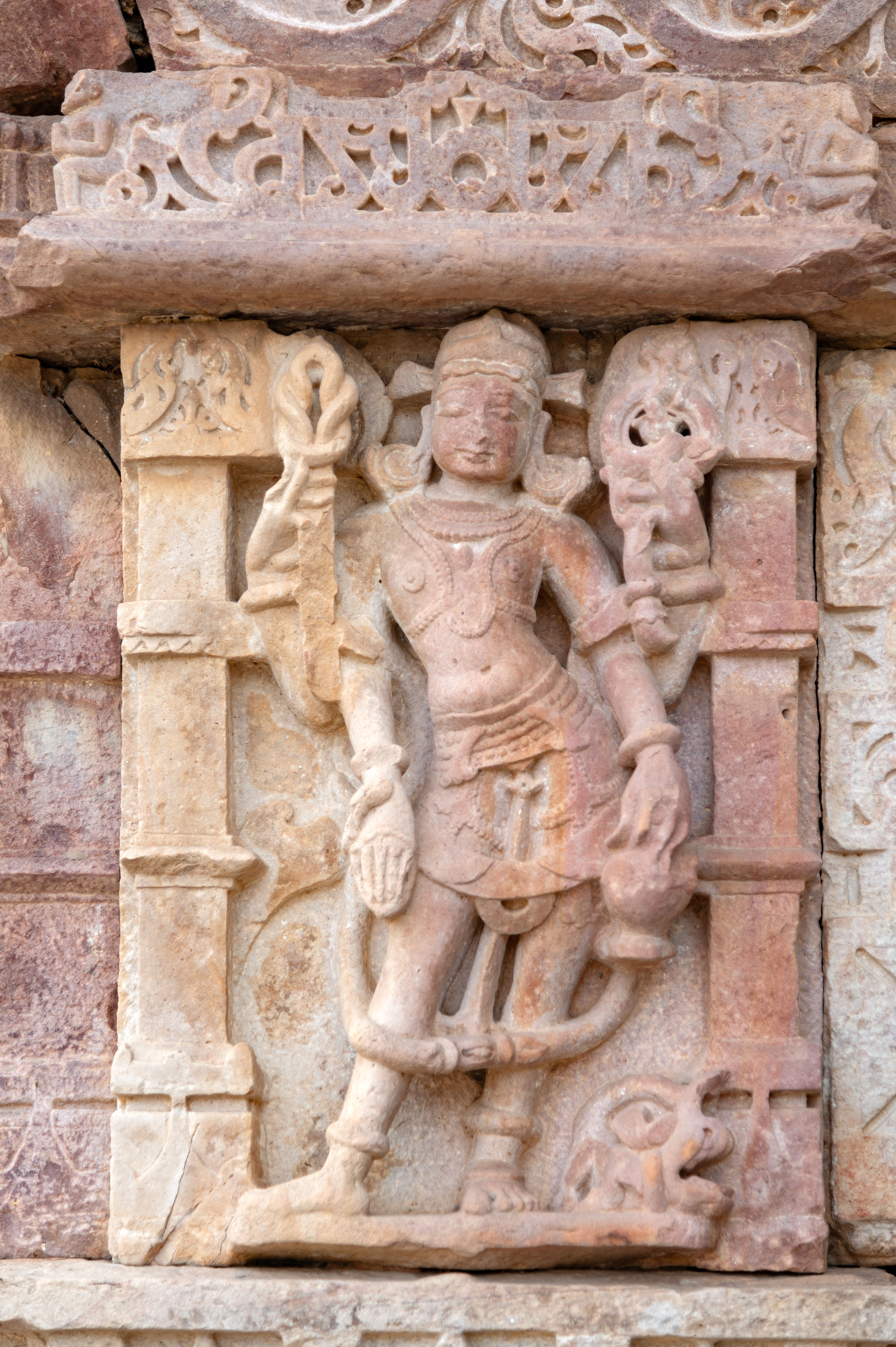 Varuna is depicted on the kakshasasna (seat back) of the Undeshwar Temple alongside his vehicle, the makara (crocodile). He holds a pasha (noose) in his upper right hand and a pushpa (flower) in his upper left hand. In his lower left hand, he carries a kamandalu (water pot), while his lower right hand is extended in the varada hasta (hand gesture of conferring a boon).