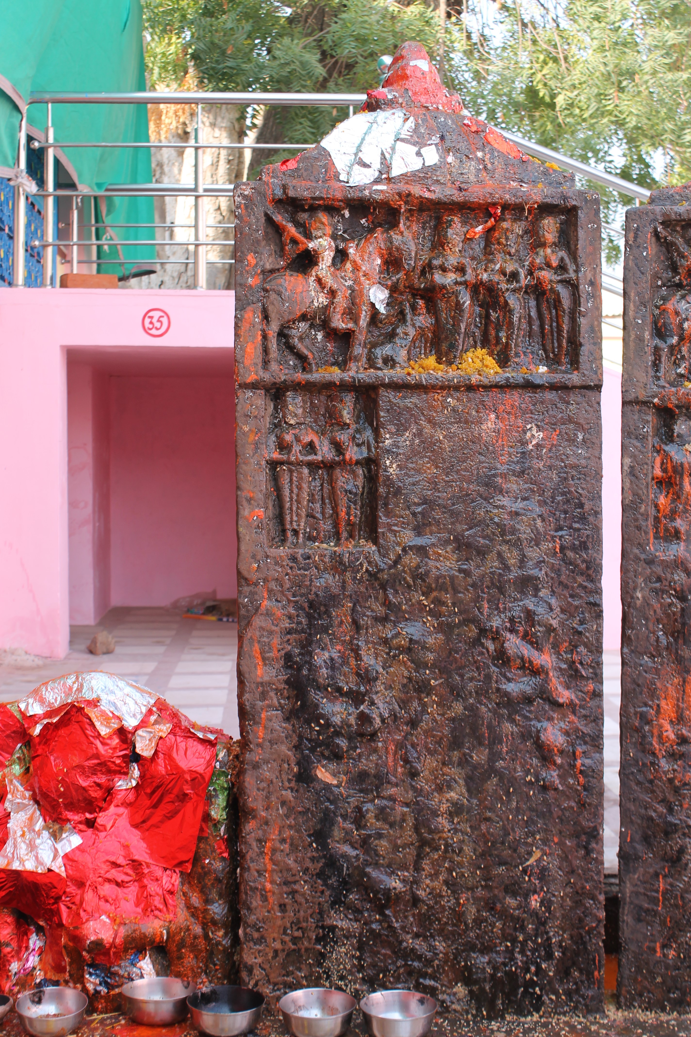 Seen here is an inscribed and sculpted hero stone (memorial stone) in the Suswani Mata temple premises, which is built in memory of the deceased soul. It is approximately five feet in height, and the front face has an inscription at the centre and human figures in the upper part. The stele is capped with a shikhara (superstructure or spire) motif.