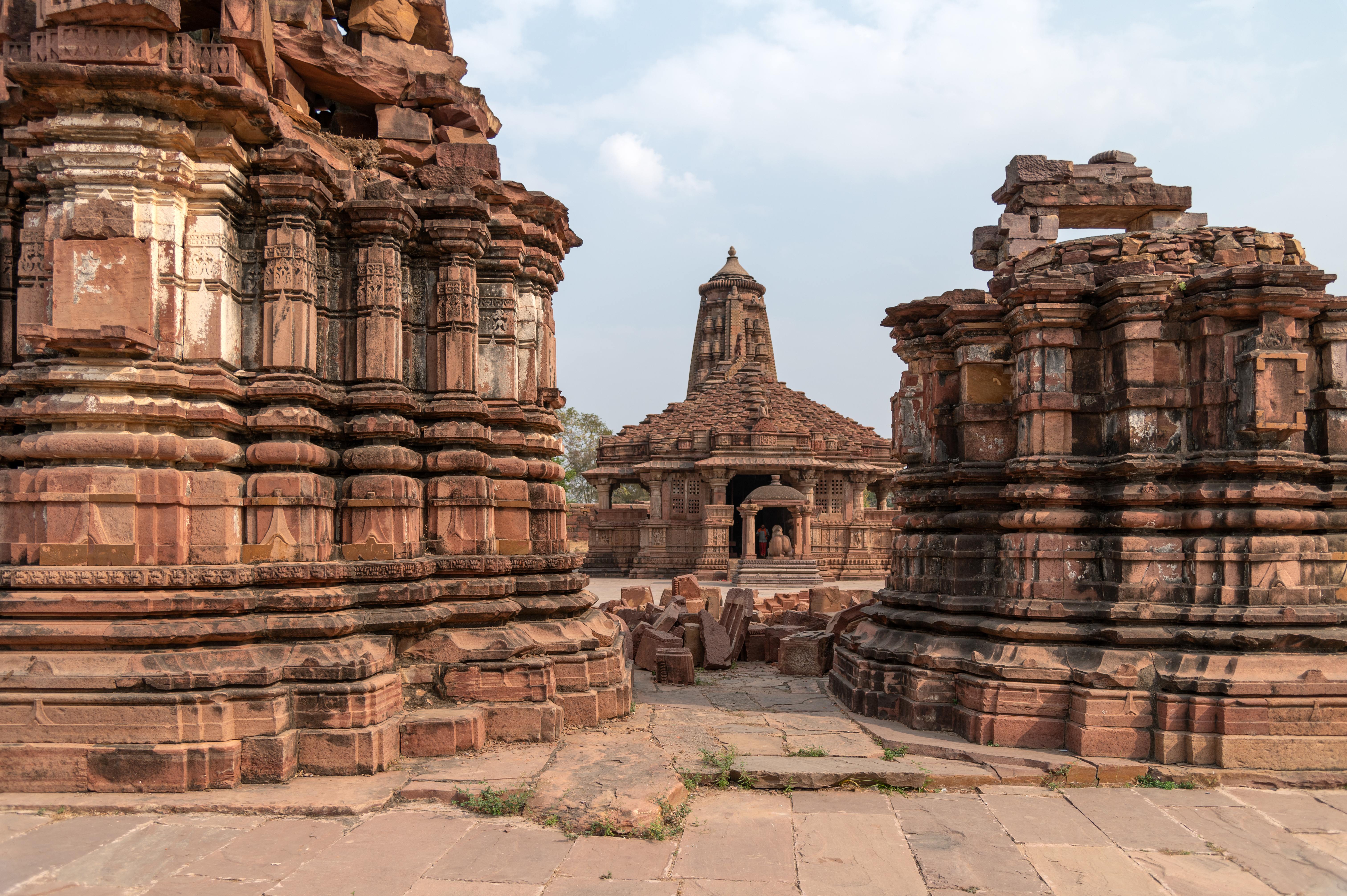 This is a view of the west-facing elevations of the two shrines that stand in front of the Mahanaleshwar Temple. These temples are built in red sandstone, consisting of plain plinth mouldings, geometric motifs on the wall subsidiary projections, and deep niches in the principal projections. The shrines have a shekhari variety of shikharas (superstructures). The walls are bereft of any icons.