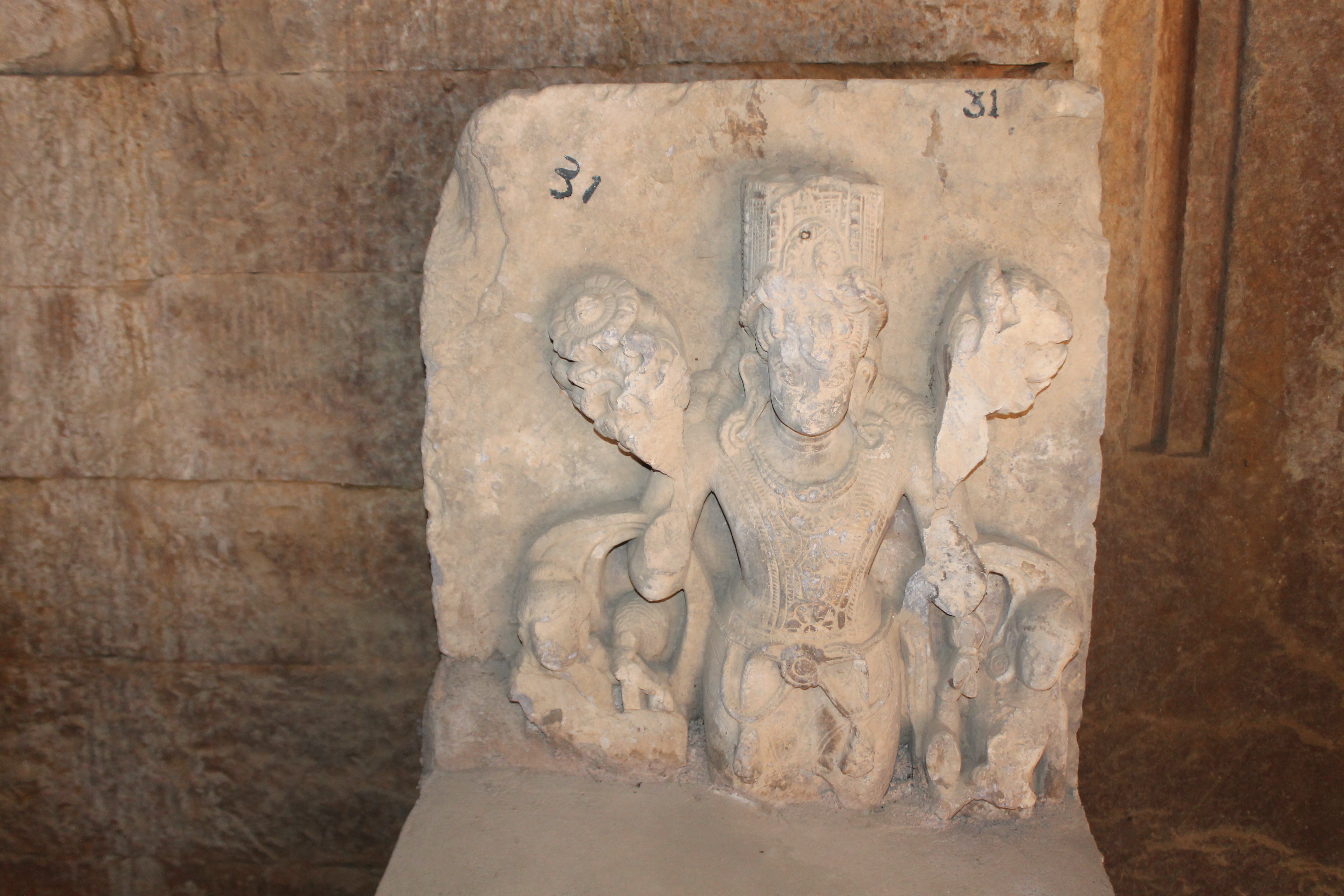 This is a broken sculpture of Surya with two lotuses in his upper hands. There are several loose sculptures placed on pedestals on the ground floor of the Mahanaleshwar temple complex's matha (monastic or residential building). The Archaeological Survey of India (ASI) collected these fragments within the Mahanaleshwar temple complex.