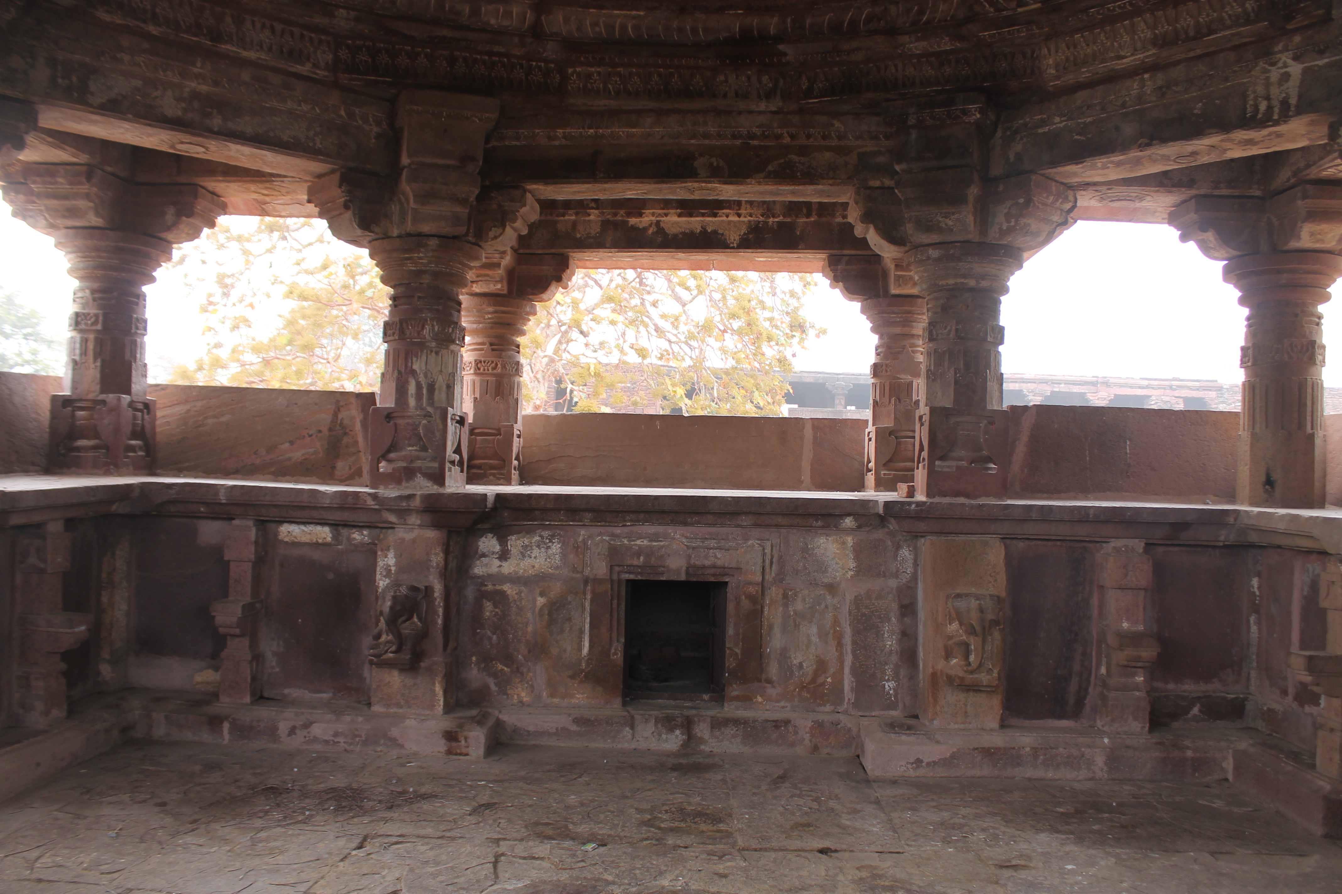 This is a view of the south-lateral transept inside the Suhaveshwar Temple's mandapa (pillared hall). The transept's dwarf wall has an opening in the center, which leads to a small room. This style of lateral wing, or transept, is similar to the mandapa of the Mahanaleshwar temple. The lower shafts of the pilasters embedded in these walls have elephant sculptures.