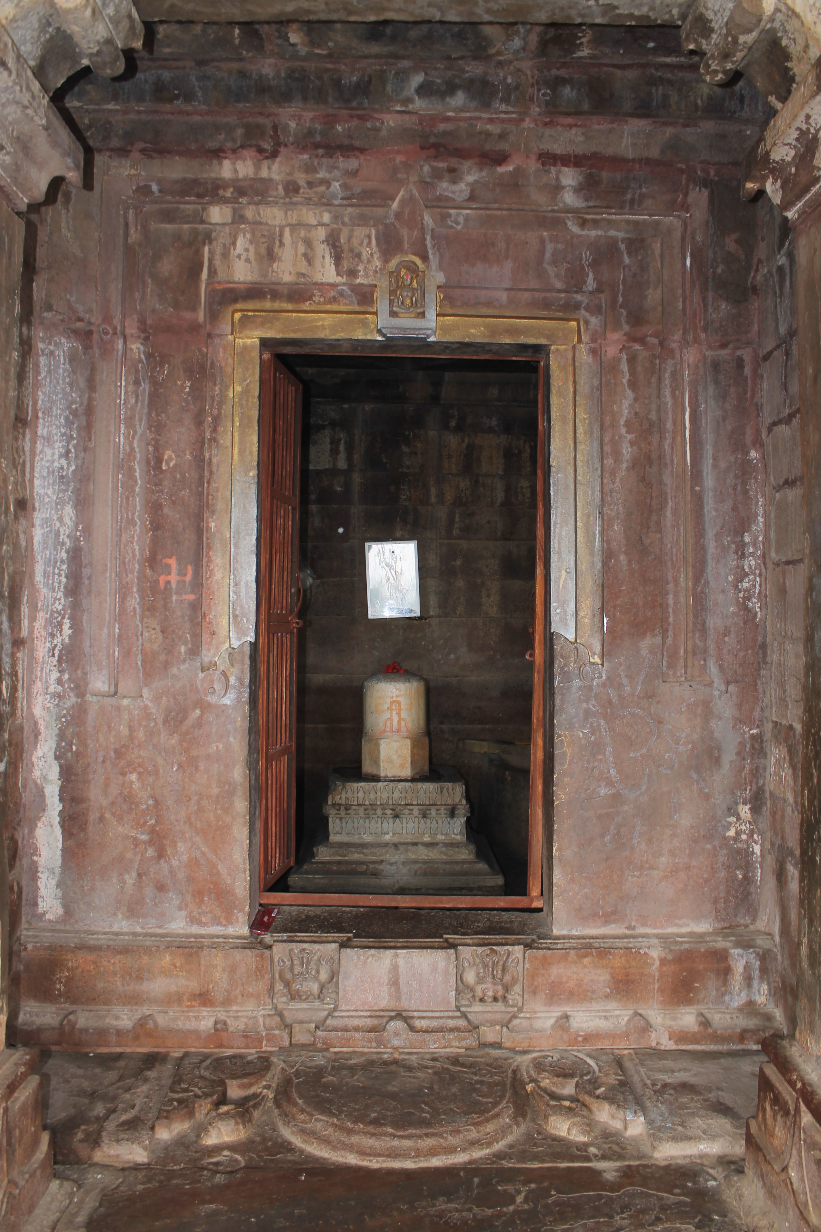 Here, you can view the details of the Suhaveshwar Temple's garbhagriha (sanctum sanctorum). The entrance is defined by an ornate dvarashakha (architrave). The pilasters flanking the entrance opening, as well as the dvarashakha's vertical and horizontal members, are all plain. Someone has painted the stone. The lintel has an icon of Lakulisa. The seated image is two-handed, holding a staff in its left hand. It is shown as nude. This sculpture on the lintel indicates that the temple was affiliated with the Pashupata tradition.