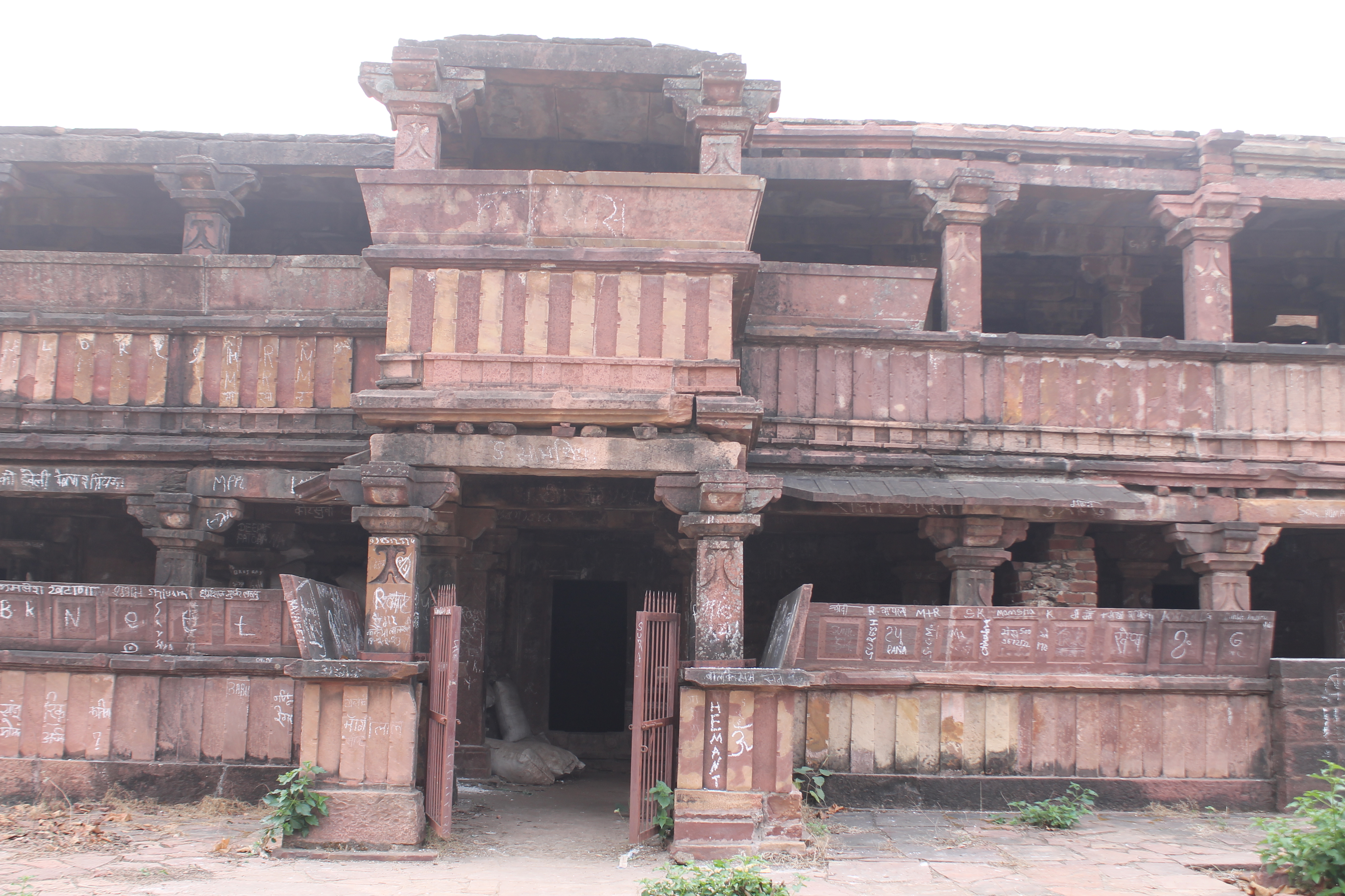 The principal entrance of the Suhaveshwar Temple's matha (monastic or residential building) is seen here, facing north. There is a protruding mukhamandapa (front porch) in the center of the façade, with a balcony above it. Both floors of the matha are semi-open. The north-facing wall is composed of dwarf walls with pillars at regular intervals. The matha's dwarf walls were once home to kakshasanas, or seat backs, but they have now lost several of these slender ones.