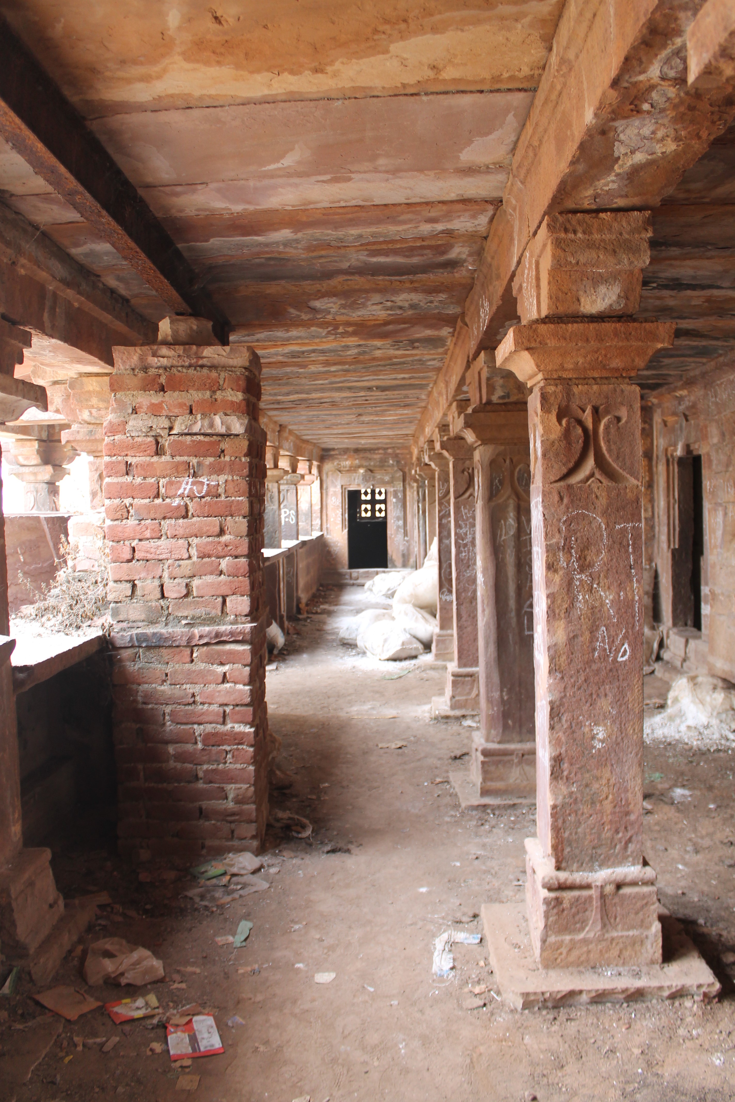 Here is a view of the colonnade on the left side of the main entrance of the Suhaveshwar Temple's matha (monastic or residential building). A row of pillars divides the space into horizontal bays. The front bay leads into two small rooms, one each at the end of the hall. The second connects to various enclosed spaces, each placed equidistantly and connected to the matha's back wall. An ornate dvarashakha (architrave) connects the enclosed rooms to the front bay.