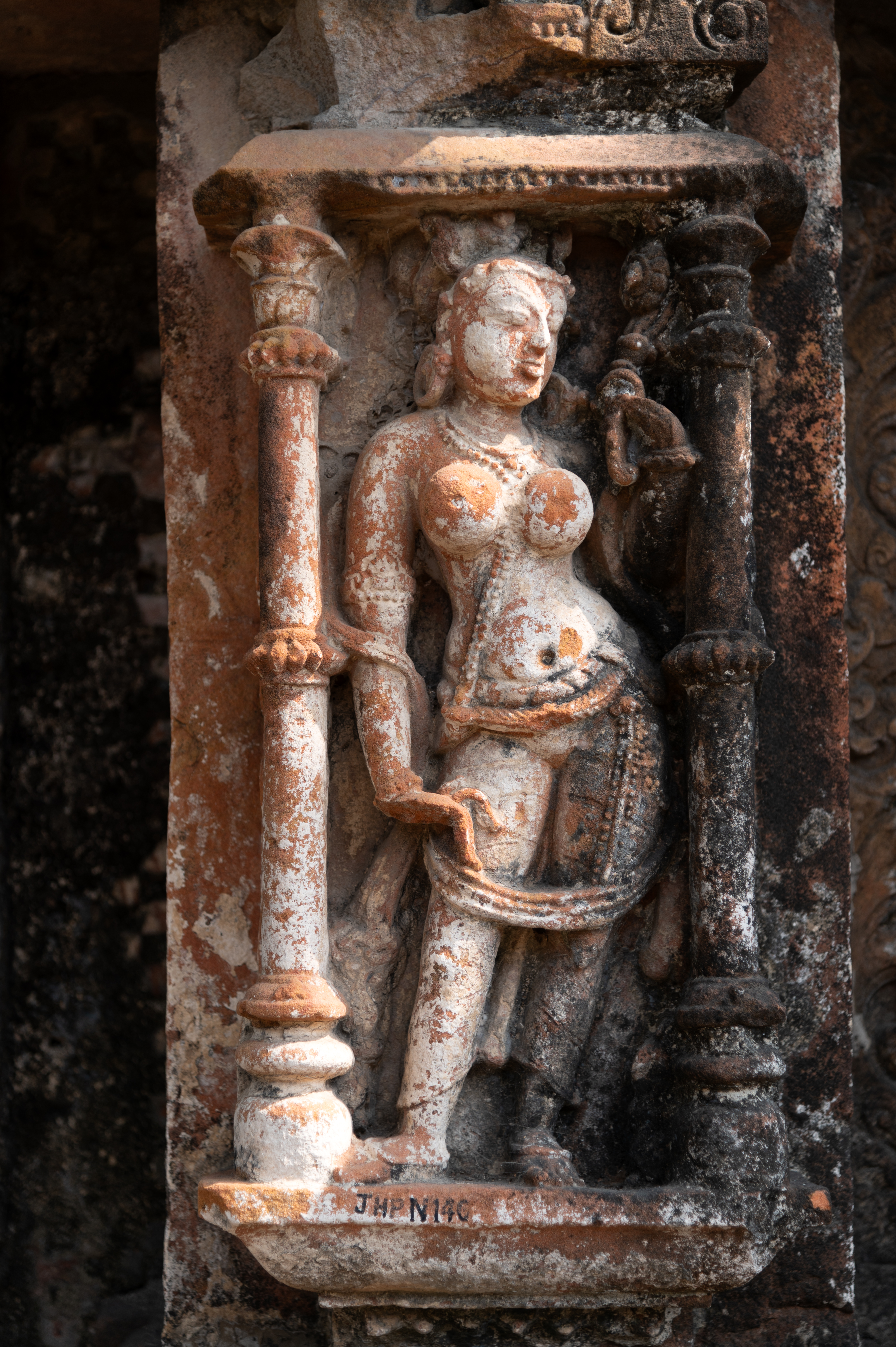 The main central niche of Shiva Temple 1 features a carved Surasundari (celestial damsel) on the west wall of the garbhagriha (sanctum sanctorum). The surasundari stands with grace, her body exuding vitality as she grasps a flower stem in one hand and the edge of a scarf in the other.