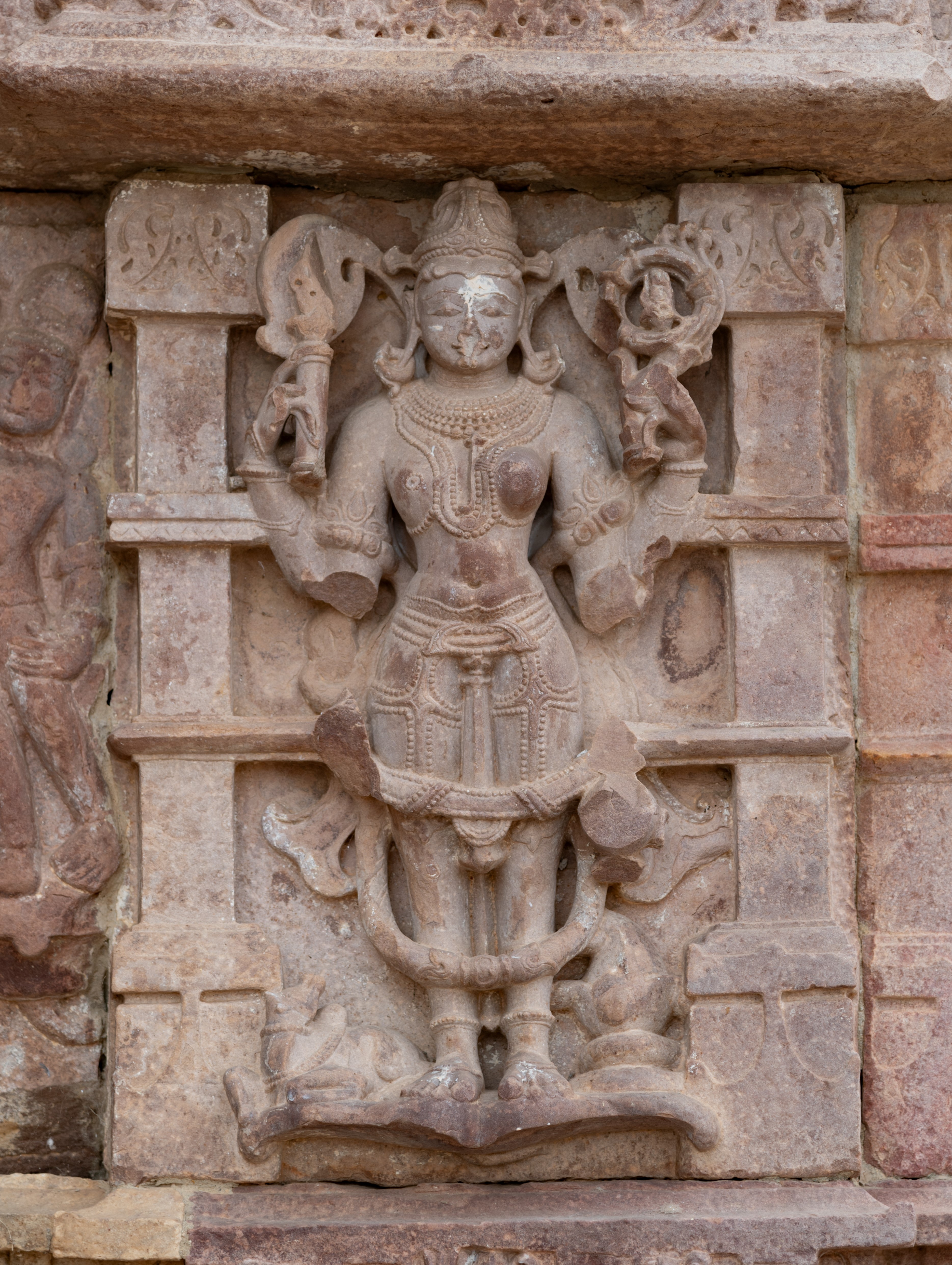  A syncretic representation adorns the kakshasana (seat backs) niche in the form of Ardhanareshwar, embodying the divine union of Shiva and Uma. Shiva’s right half and Uma’s left half seamlessly blend into a single divine form, symbolizing the inseparable nature of masculine and feminine energies. While the figure originally had four arms, only the upper two remain intact. Shiva’s side wields a pointed weapon, signifying his power, while Uma’s side holds a pushpa (flower), representing her nurturing aspect. At the bottom, Nandi, Shiva’s mount, accompanies his side, while Uma’s side features a kalasha (pitcher) adorned with verdant vegetation, symbolizing fertility and abundance. It is significant because both the syncretic images of Harihara and Uma-Maheshvara connect Vishnu and Uma with vegetal growth, symbolizing prakriti (nature), while Shiva represents purusha (essence).