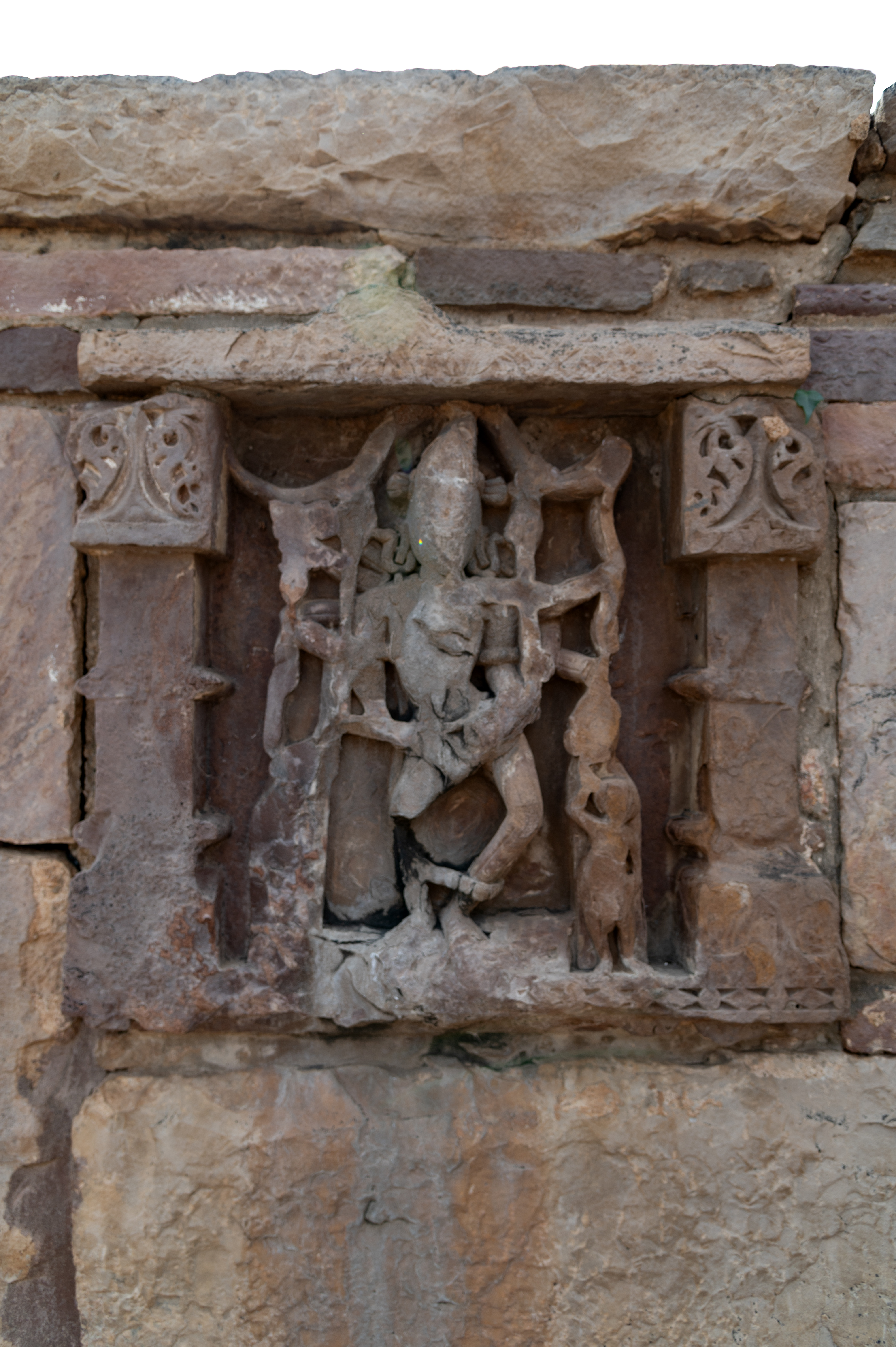 Image 14: Within the enclosure wall is a depiction of Chamunda, portrayed with multiple arms. Although the image has faded over time, some characteristics are still discernible. Chamunda is depicted holding a sarpa (snake) in her upper arms, a khatvanga (staff) in one hand, and a severed human head in another. Additionally, a human figure is shown to her left, attending to her.