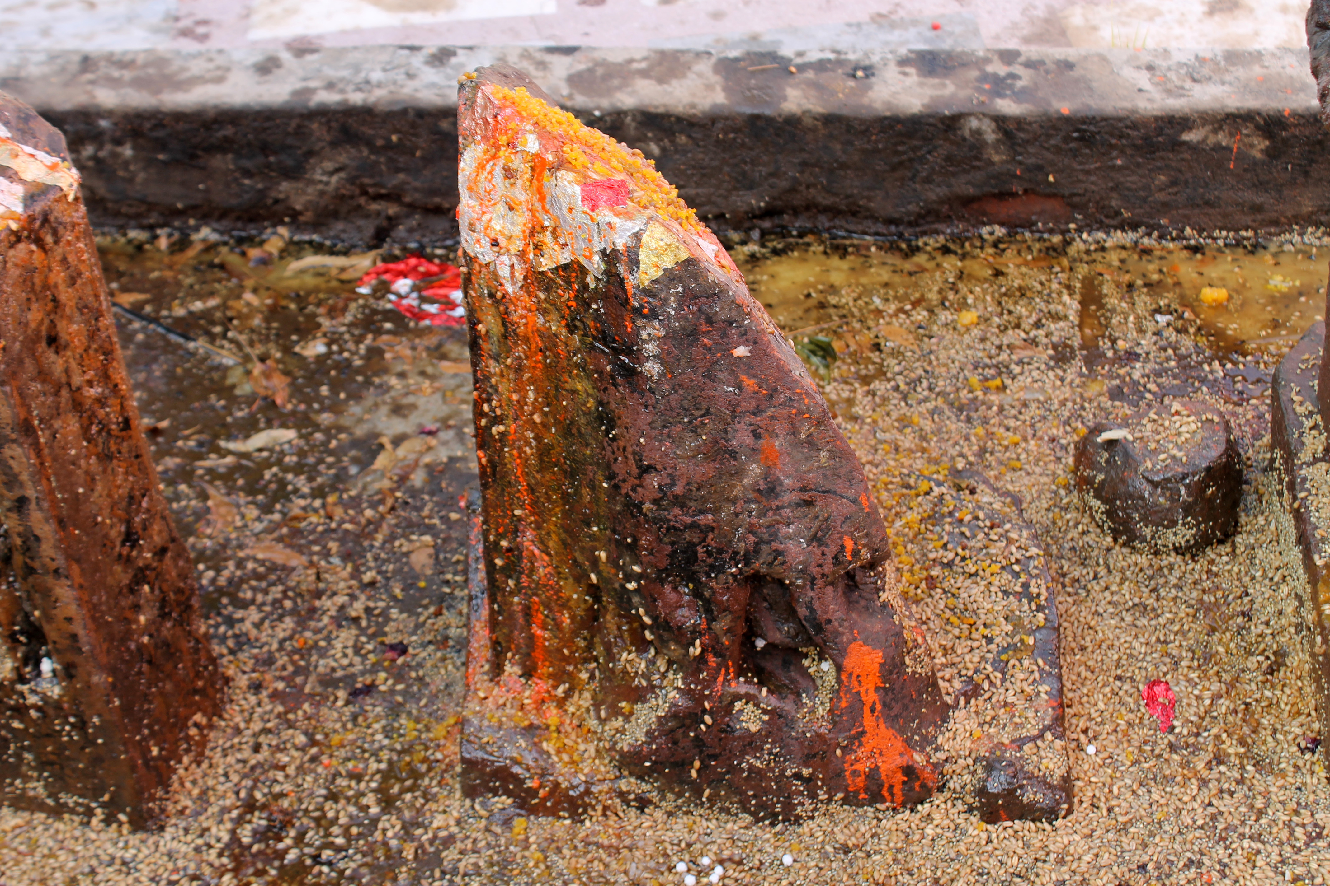 Seen here what appears to be a sculptural fragment among the row of hero stones. This speculation is drawn from faint traces of a human figure visible on the stone fragment. Given that the collection includes not only hero stones but also other pieces, it is plausible that a broken sculpture is venerated as a hero stone. Unlike other Hero stones, this fragment has a low pedestal.
