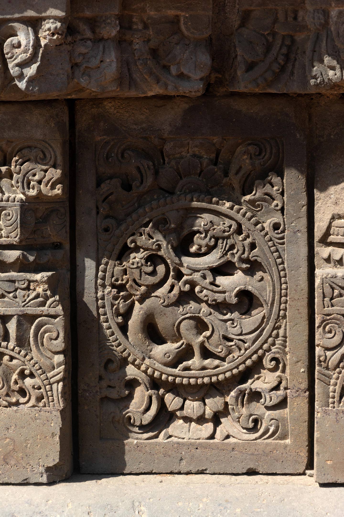 The plaque's central artwork consists of a circular medallion surrounded by a foliage motif. A bird, likely a peacock, with its head bent downward, integrates with a creeper motif in this medallion. The rest of the plaque is covered with ardha padma (half lotus) and kalpa lata (creeper) motifs.