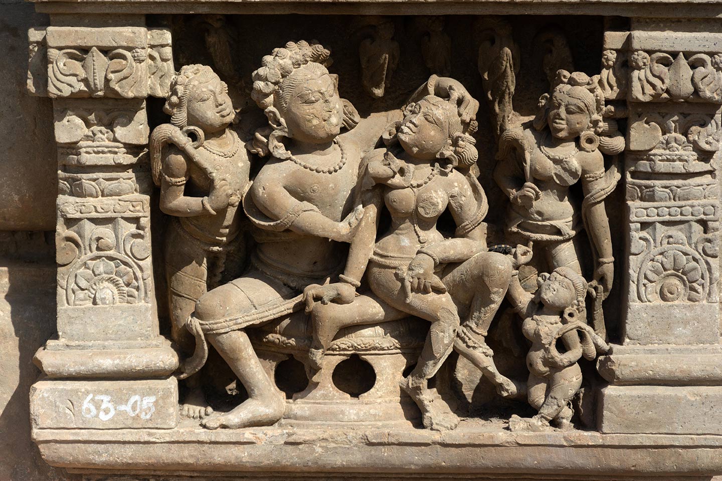 Panels with pillars and tiered shikhara featuring gavaksha (horseshoe) motifs frame couples invoking shringara rasa, which is associated with romance, love, and attractiveness between lovers. The central male figure is seated in the lalitasana pose on a raised seat. The central female figure sits on his left thigh, her head turned towards the man. The man is admiring the beauty of the woman, placing his left hand on her coiffure and inspecting her face with his missing left hand.