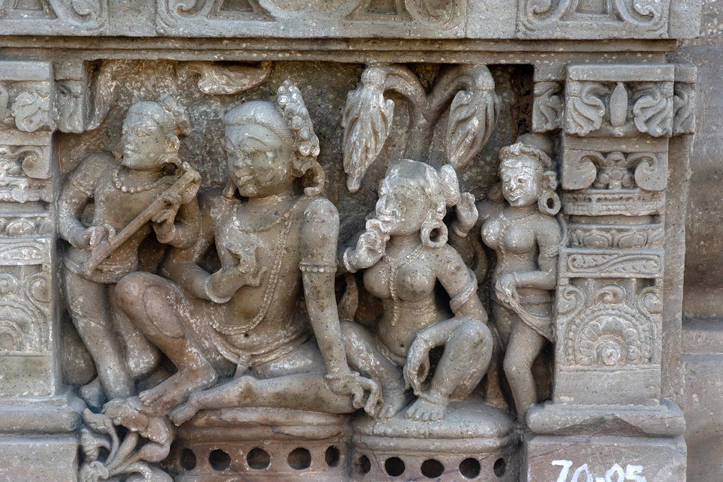 The central male figure is seated in the rajalilasana pose (both legs raised on the seat, palms of the feet together) on a raised seat. In the malasana (squat) pose, the central female figure is seated on the left of the man, in a separate seat. Male and female attendants flank the central figure from the left and right, respectively. All the figures are watching a performance (not in the frame) and looking in unison towards the left.