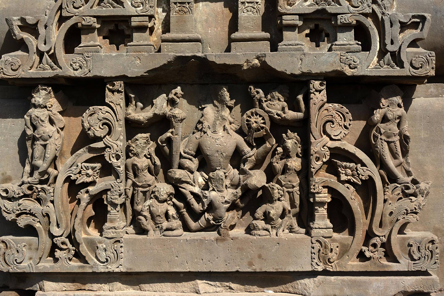 Image 3: The depiction of Lord Vishnu (Aniruddha) is chaturbahu, meaning he has four hands. He carries the gada (mace), named Kaumodaki, in his rear right hand, and the chakra (discus), named Sudarshana, in his rear left hand, while his front two hands remain broken. On the top corners, we see a pair of vidhyadharas (knowledge bearers), showering floral garlands.