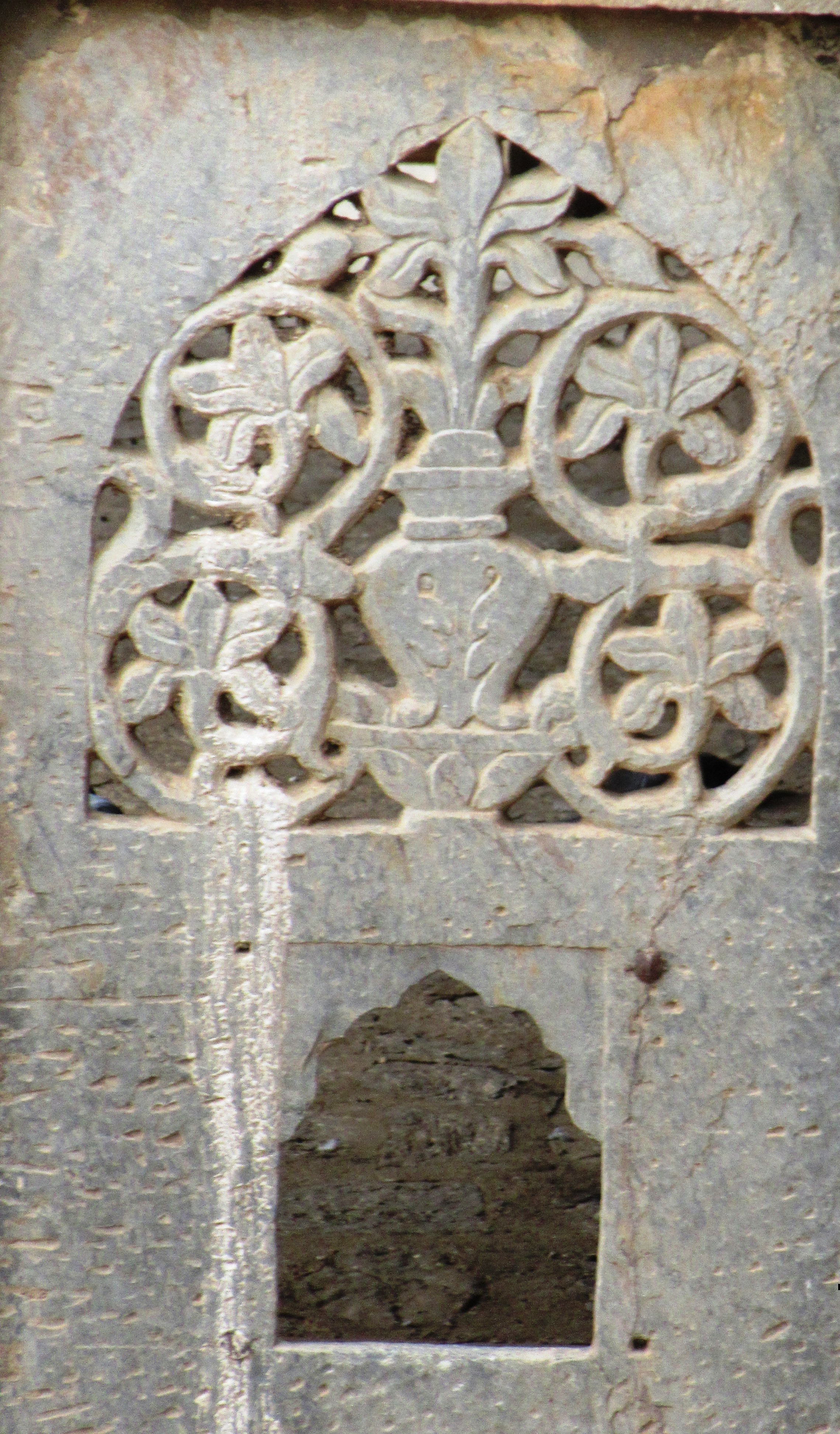 Image 2: A lattice window showing a flower vase. The room is colonnaded and spacious, complete with cusped arches. Two flights of stairs on both ends allow entry into the 'palace'. The arcade is four-sided with pointed arches. It is also known as the Summer Palace. Small jalis are present on the windows.