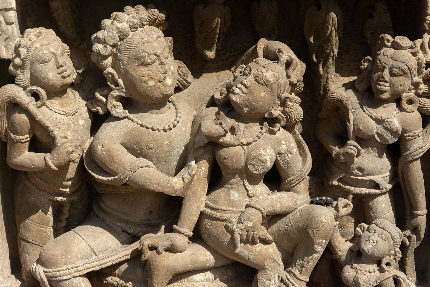 Image 5: The central male figure is seated in a lalitasana pose on a raised seat. The central female figure is seated on his left thigh, looking towards the man with her head turned. The man is admiring the beauty of the woman, placing his left hand on her coiffure and inspecting her face with his left hand (missing). Chauri dharini (fly whisk bearers) stand on either side of the couple, looking towards them. A small female figure features at the right bottom, likely an attendant or servant.